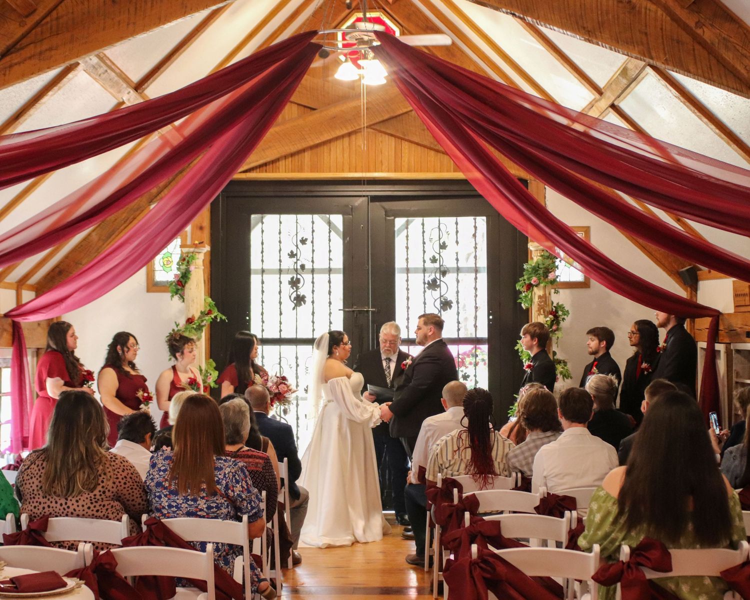 Indoor wedding ceremony in a wooden chapel with draped red fabric and stained glass windows during a traditional service.