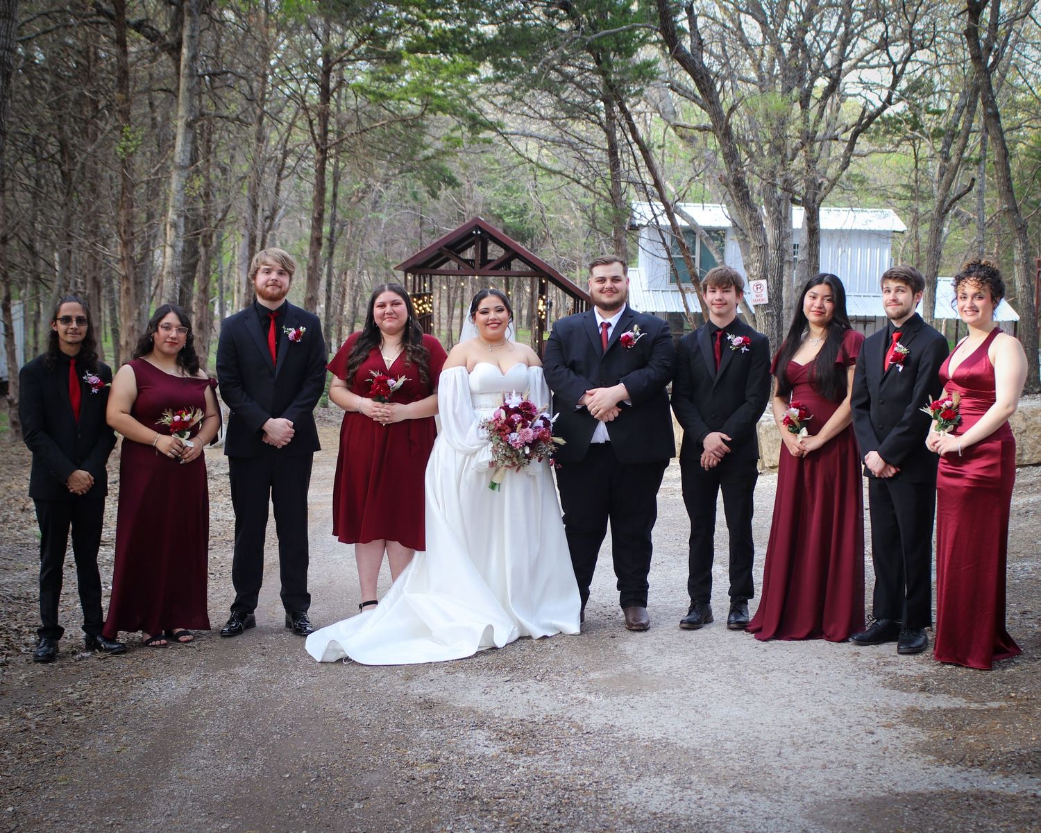 Wedding party in burgundy dresses and black suits pose together on a tree-lined pathway.