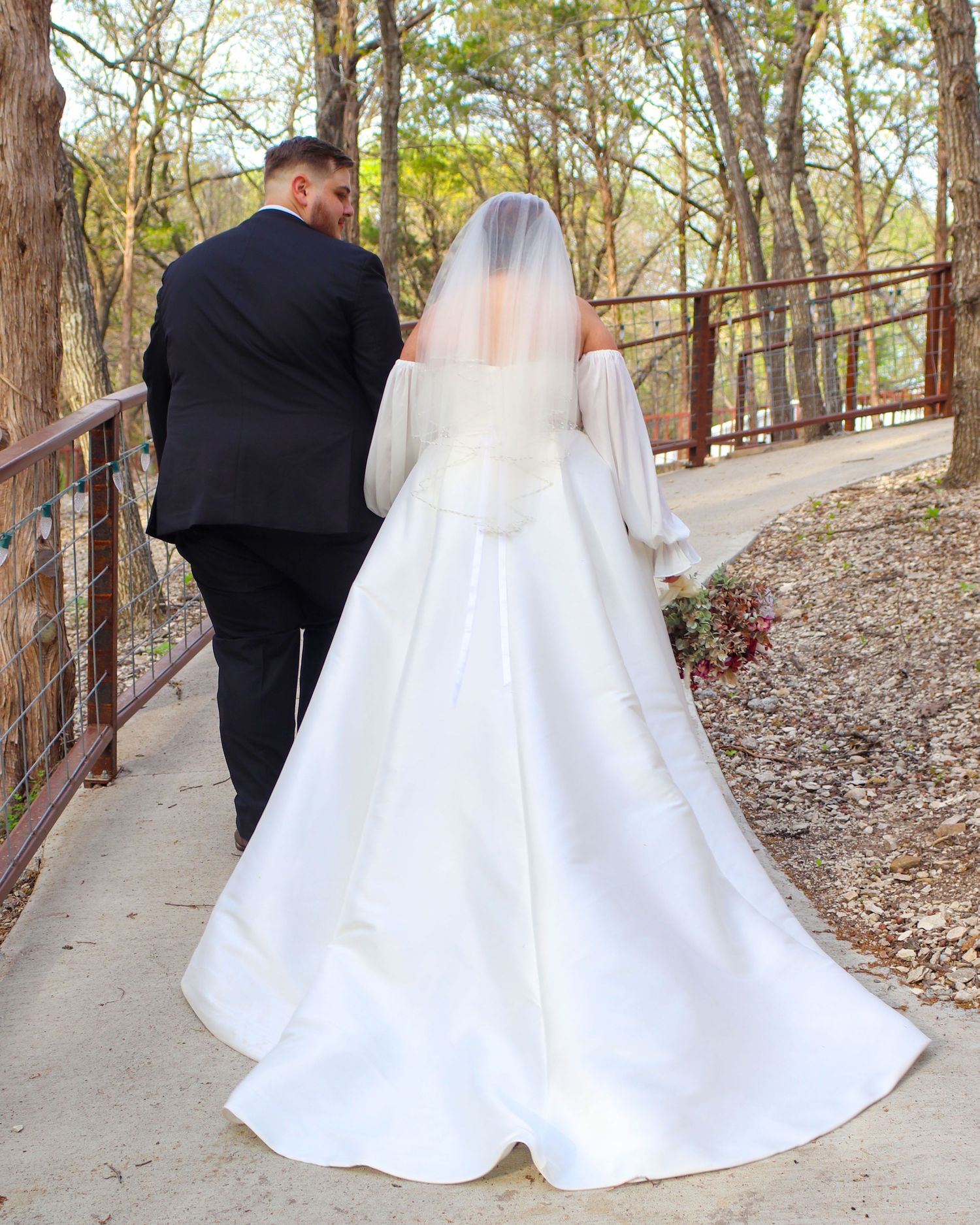 A couple wearing a black suit and white wedding dress walk along a wooded pathway with a rustic metal railing.