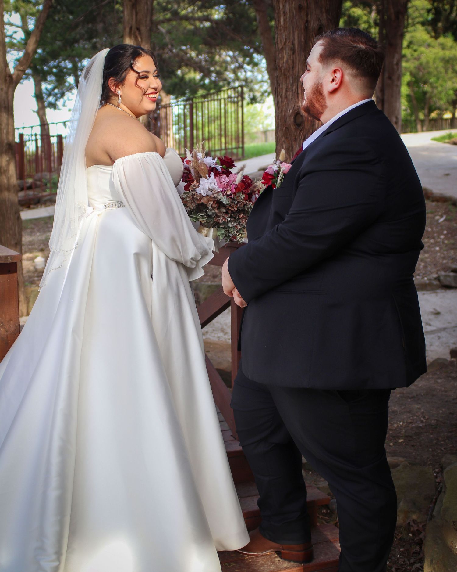 A wedding couple shares a joyful moment outdoors with the person in white holding a vibrant purple and red bouquet.