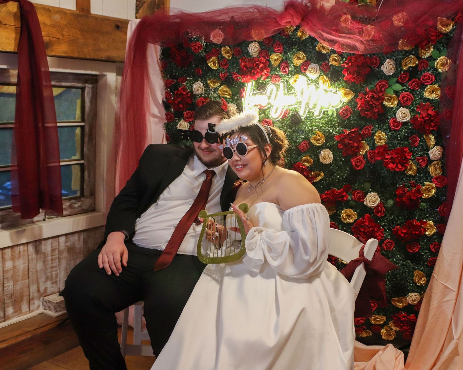 A couple in wedding attire poses on a couch in front of a red rose wall with neon lighting.