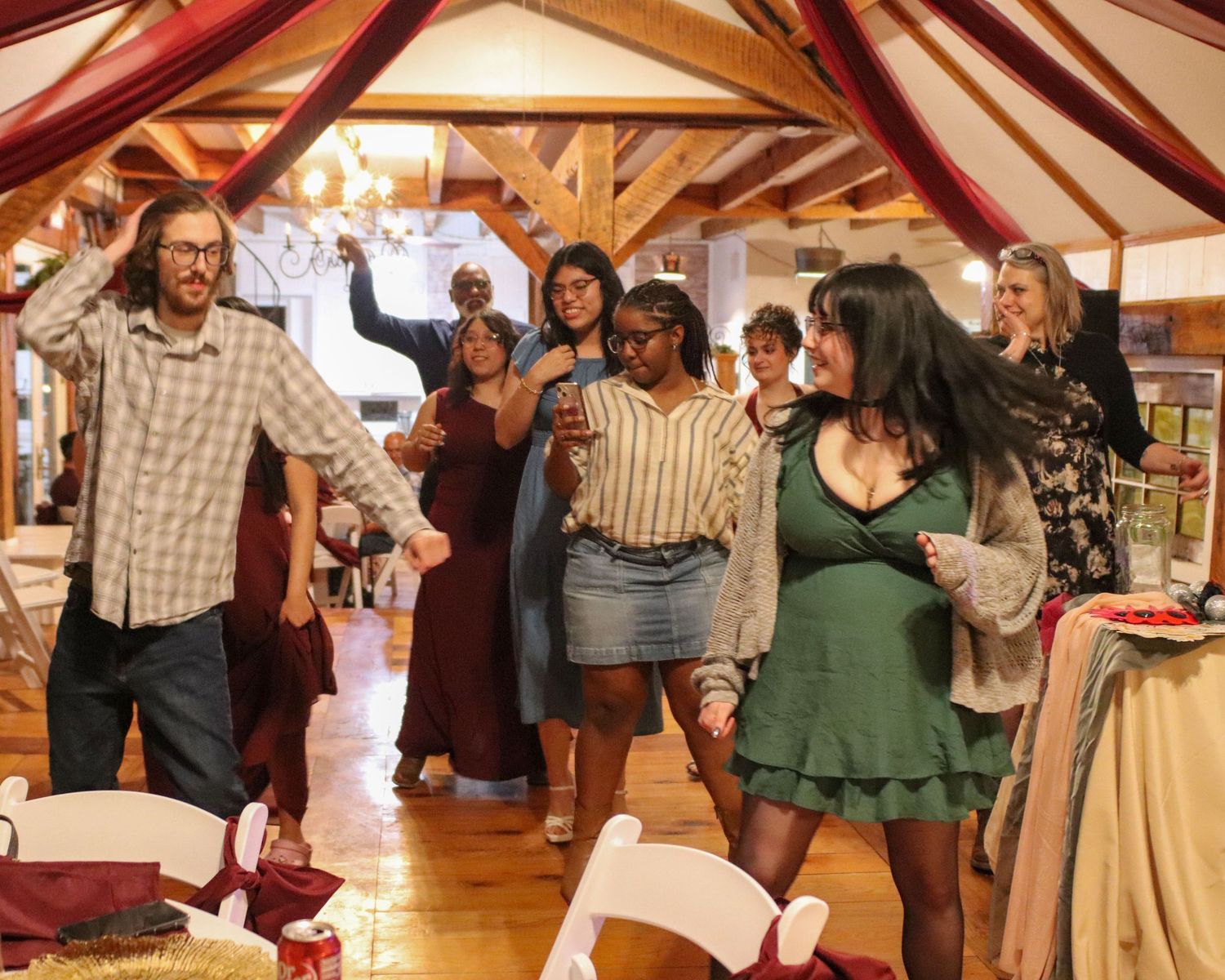 Wedding reception guests dance and celebrate in a rustic tent-draped venue with wooden beams.