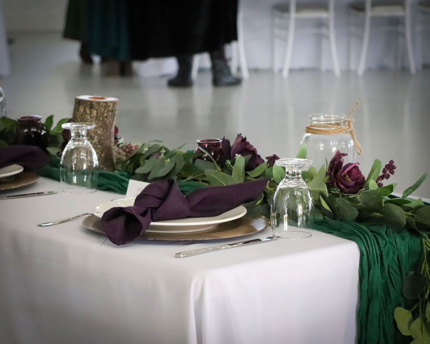 Elegant table setting with purple napkins, green foliage and glass candle holders.