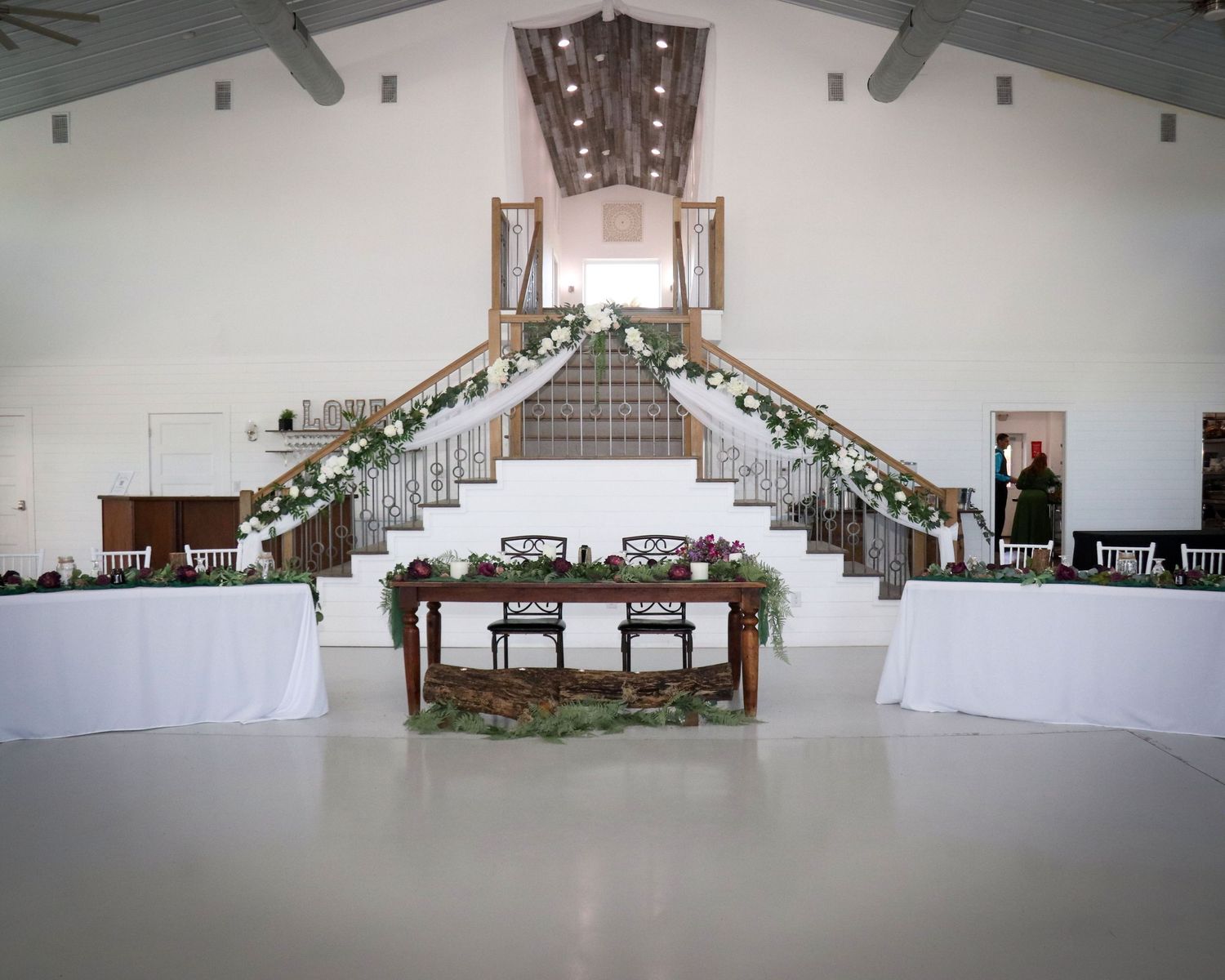 An elegant wedding reception venue with a grand staircase decorated with greenery garlands and white linens at dining tables.