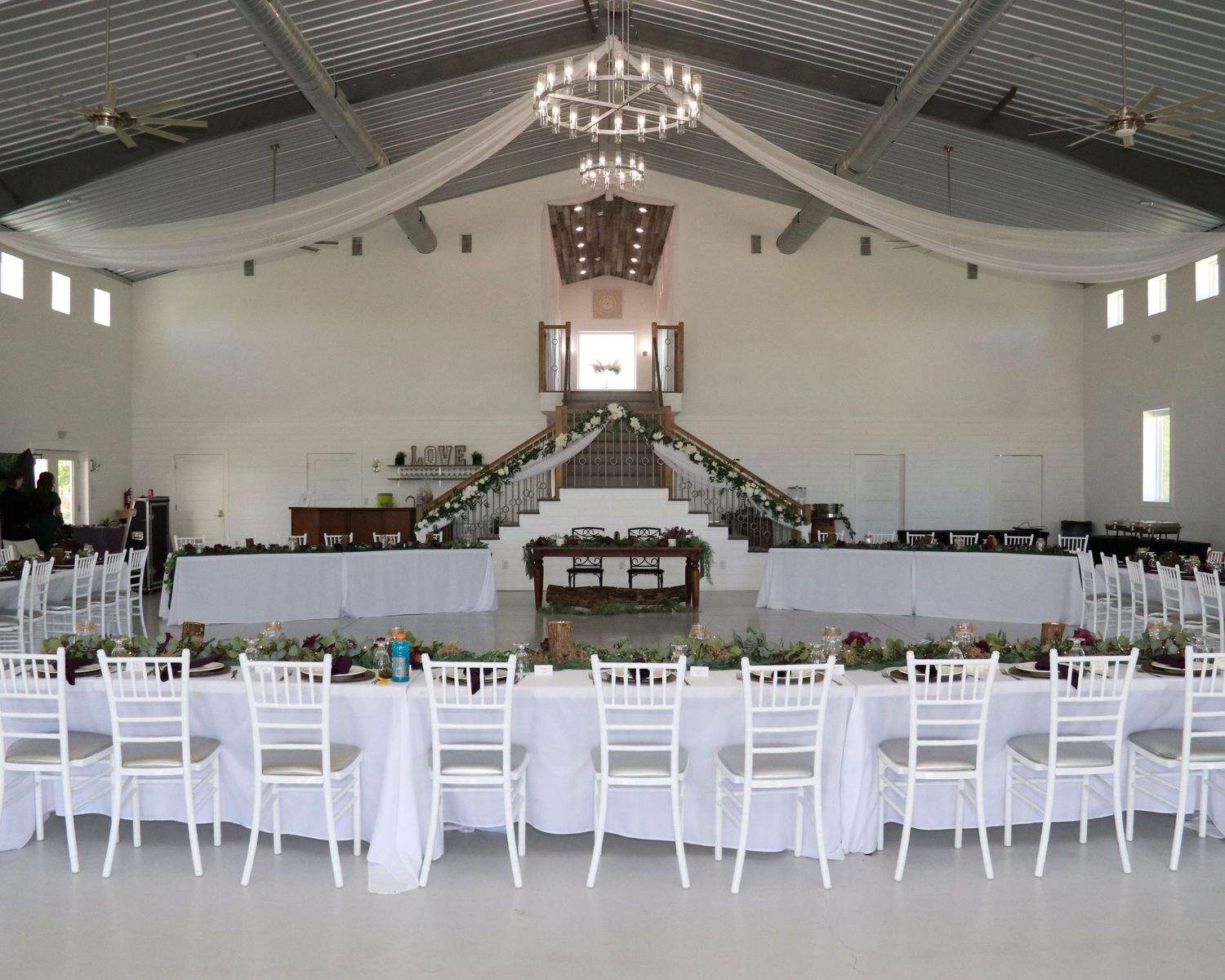 Elegant white wedding reception hall with chandelier, draped fabric, and rows of white chairs set for a celebration.