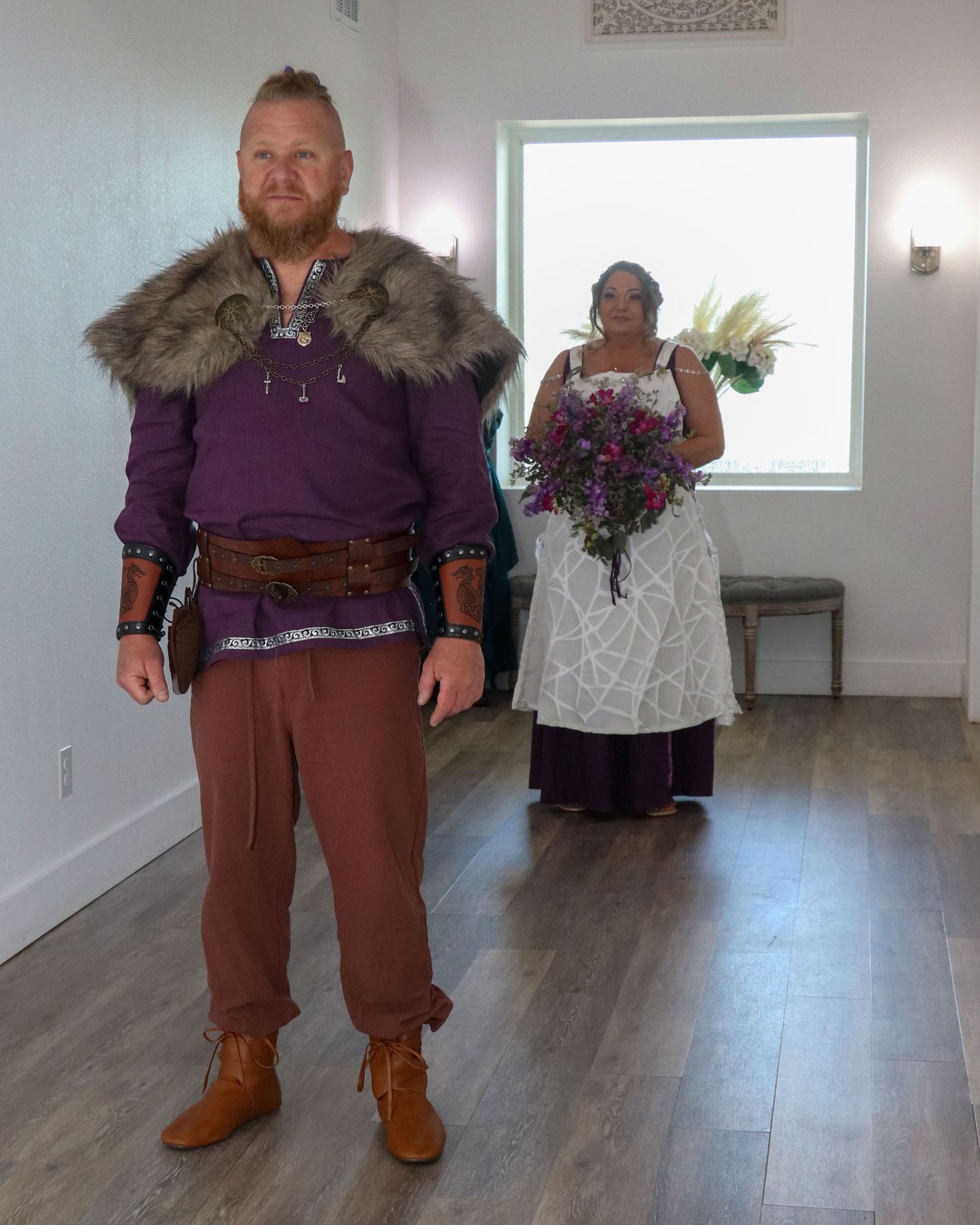 A couple in medieval-inspired wedding attire pose together in a bright indoor space.
