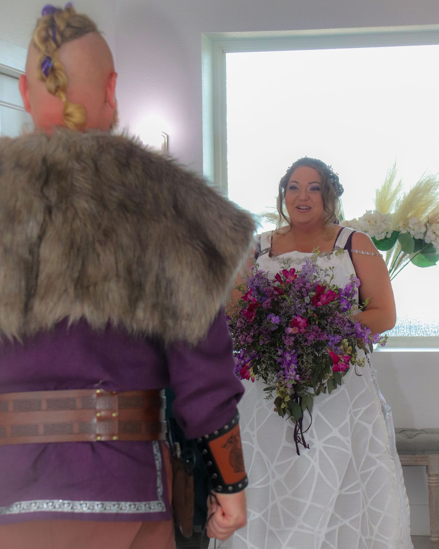 Couple in wedding attire featuring a purple viking costume and white geometric dress with floral bouquet.