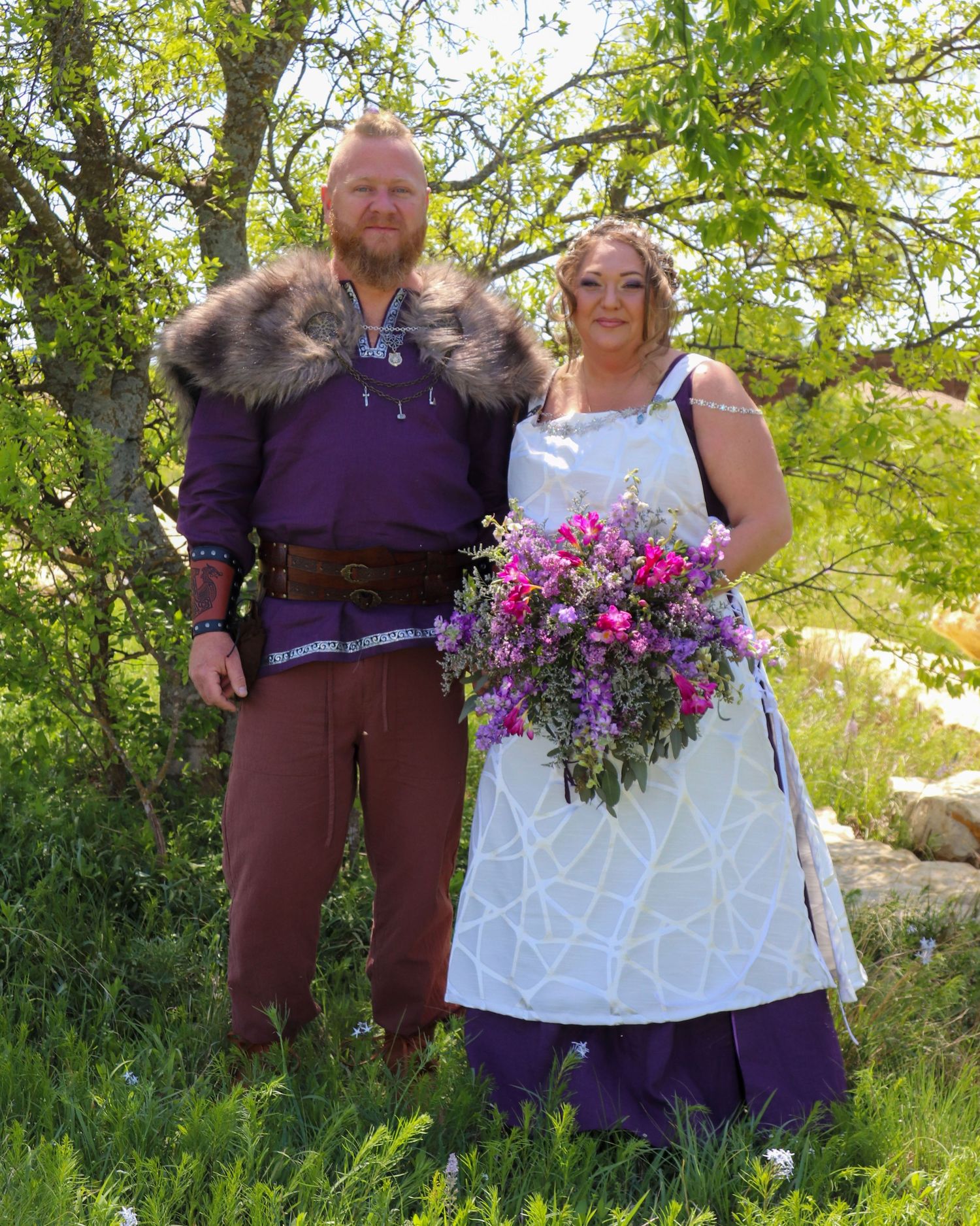 Wedding couple in viking and medieval inspired outfits pose outdoors among green foliage.
