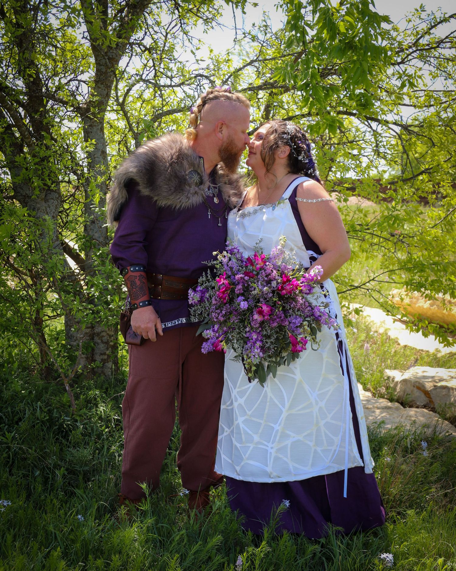 A couple shares a tender moment in a lush garden setting with a vibrant purple and pink floral bouquet.