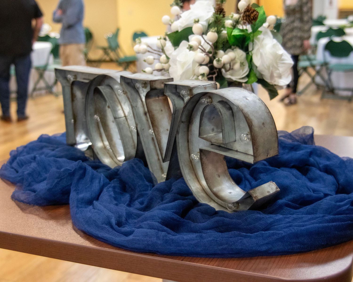 Silver "Love" decorative letters arranged on blue fabric with white floral accents on a wooden table.