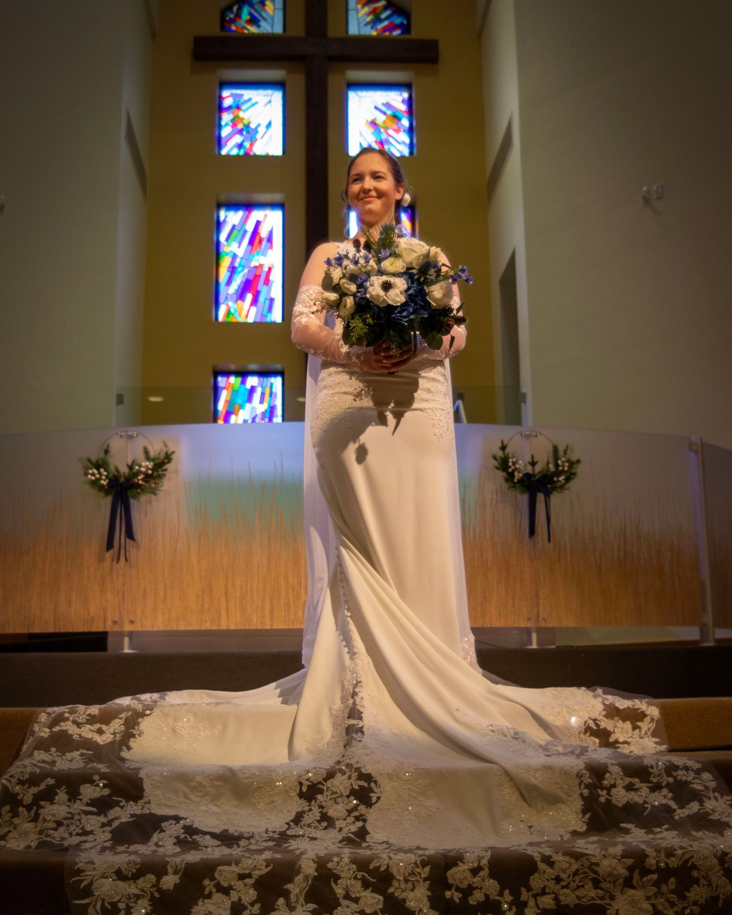 A bridal figure stands in a flowing white gown against stained glass windows in a church setting.