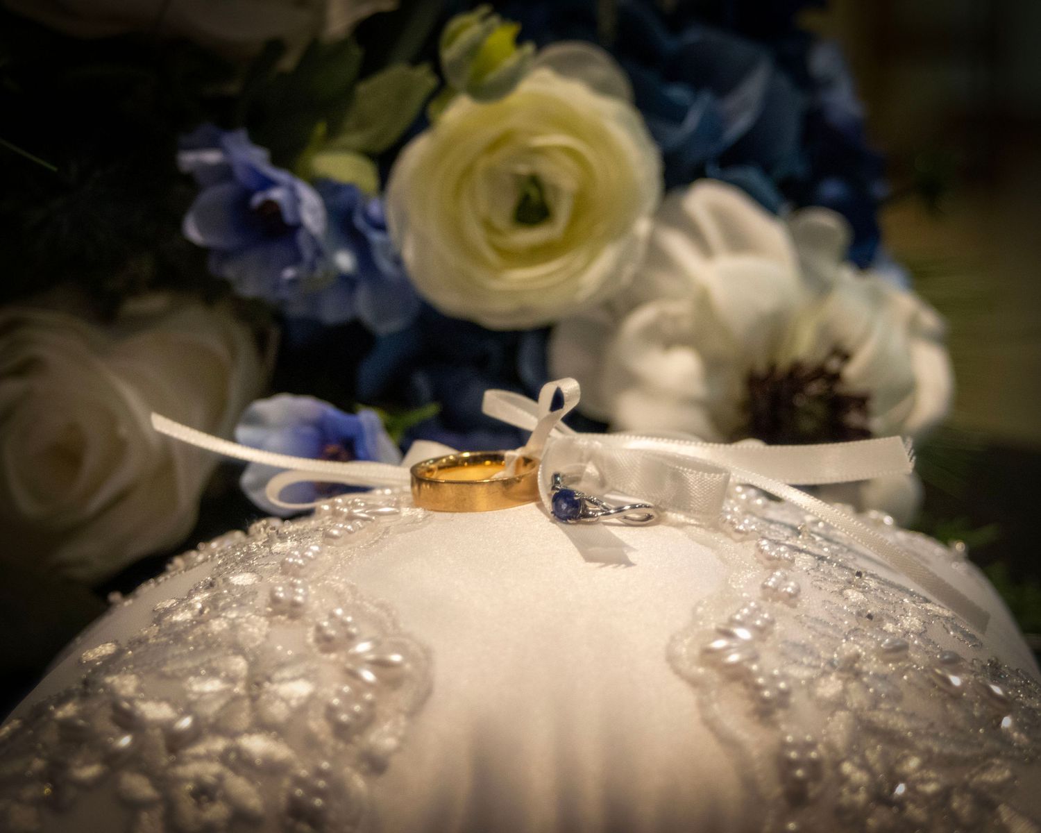 Wedding rings resting on a white lace wedding dress with blue and cream flowers in the background.
