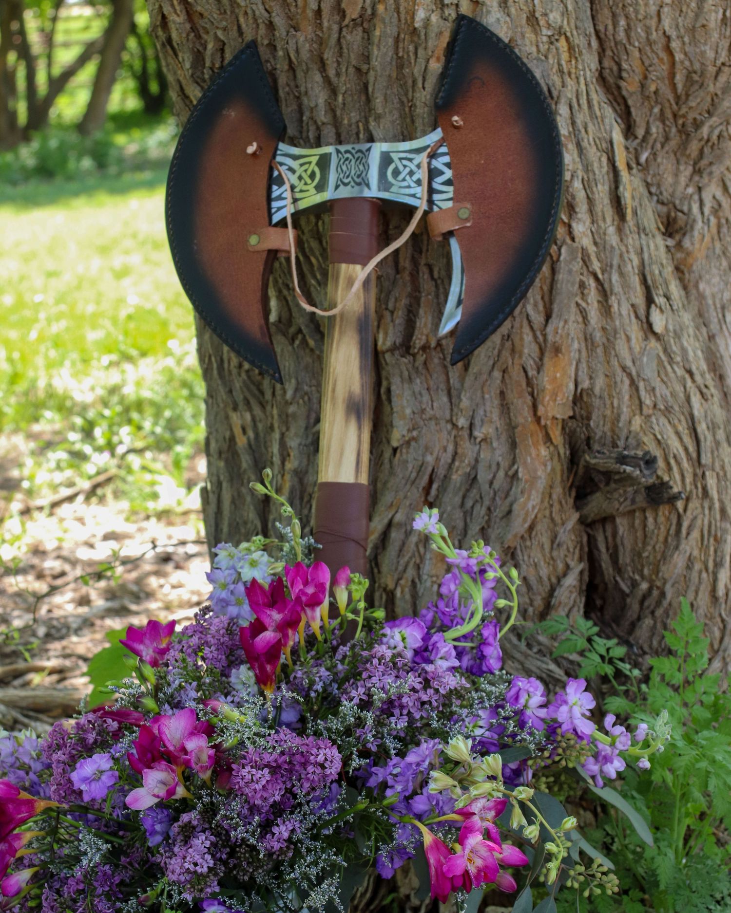 Medieval style double-headed axe displayed on tree trunk with vibrant purple and pink floral arrangement.