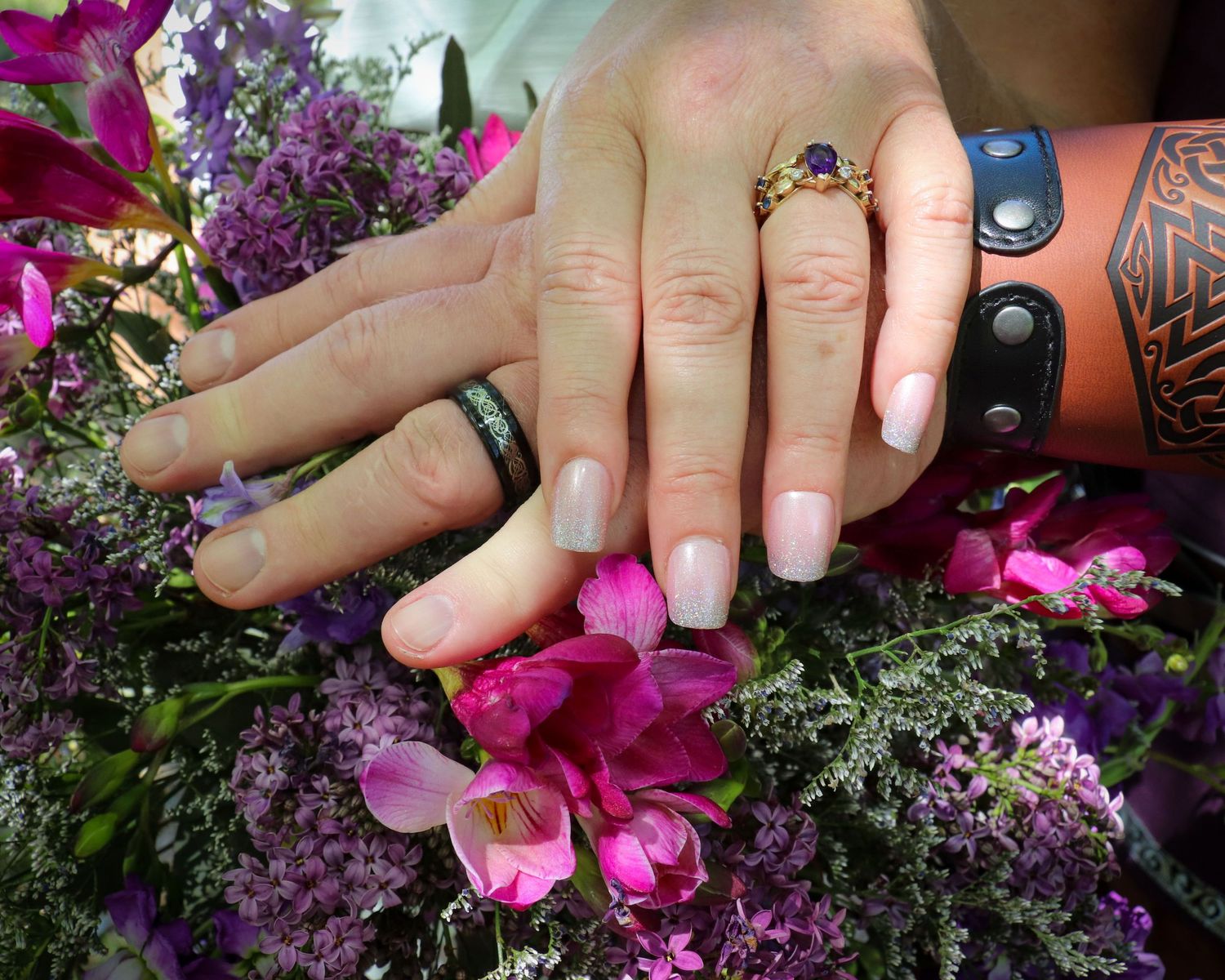 Close-up of hands with wedding rings resting on vibrant purple and pink flowers.