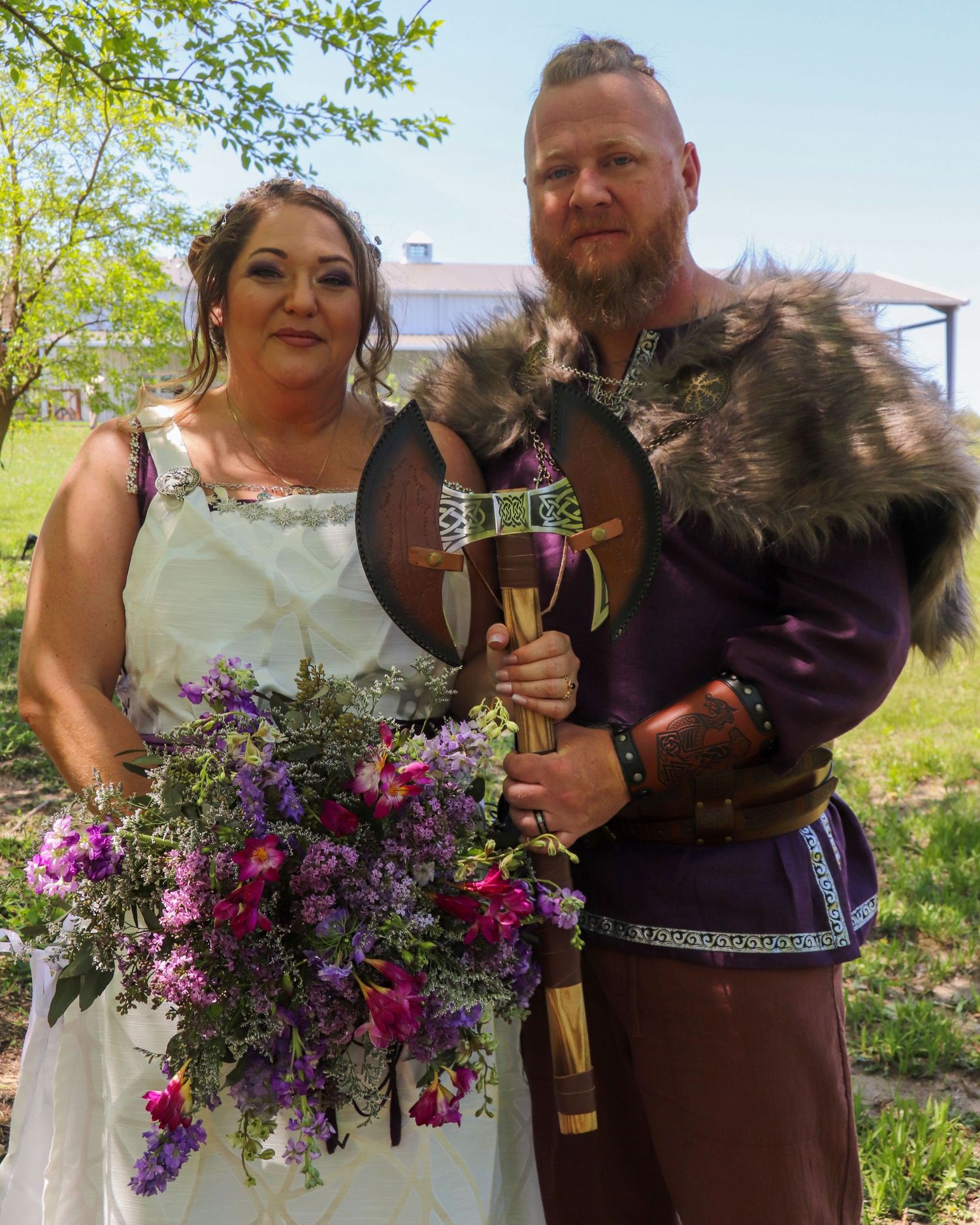 Viking style wedding couple in traditional dress with purple floral bouquet and medieval attire stand together outdoors.