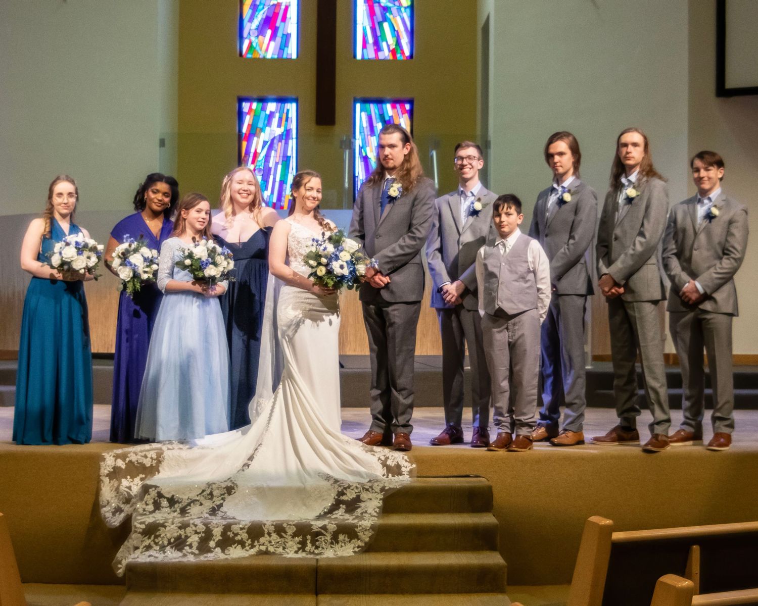 Wedding party stands on church altar steps with stained glass windows and floral arrangements in the background.
