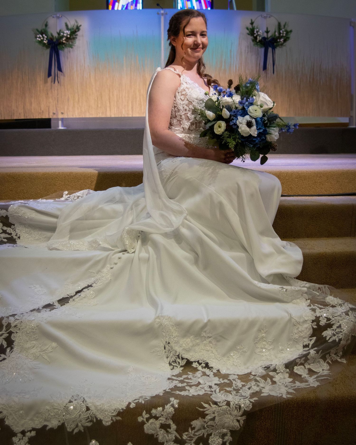 A bride in a flowing white gown sits on church steps with a blue and white floral bouquet against a light-colored backdrop.