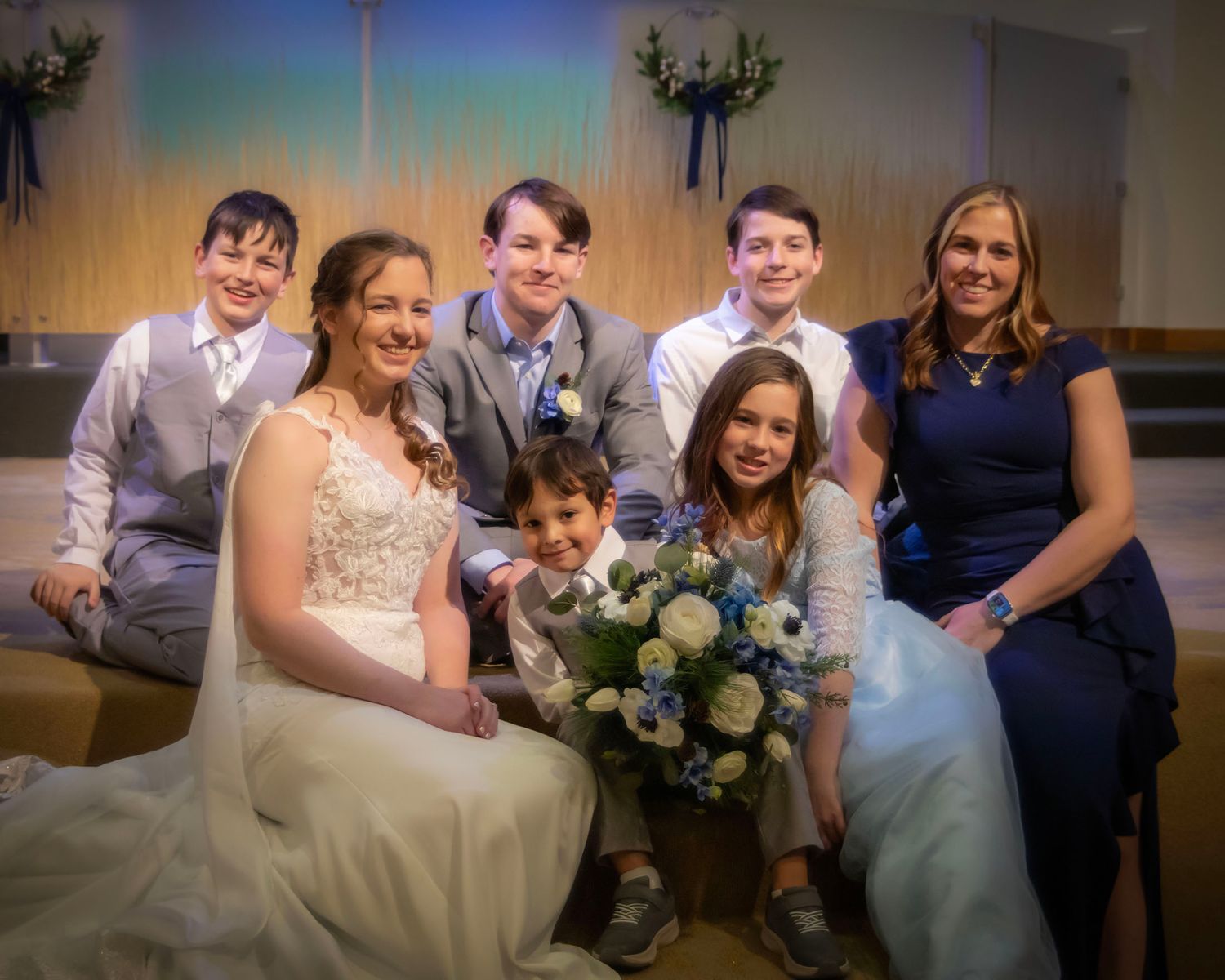 A wedding party poses together with a floral bouquet in a softly lit indoor venue.