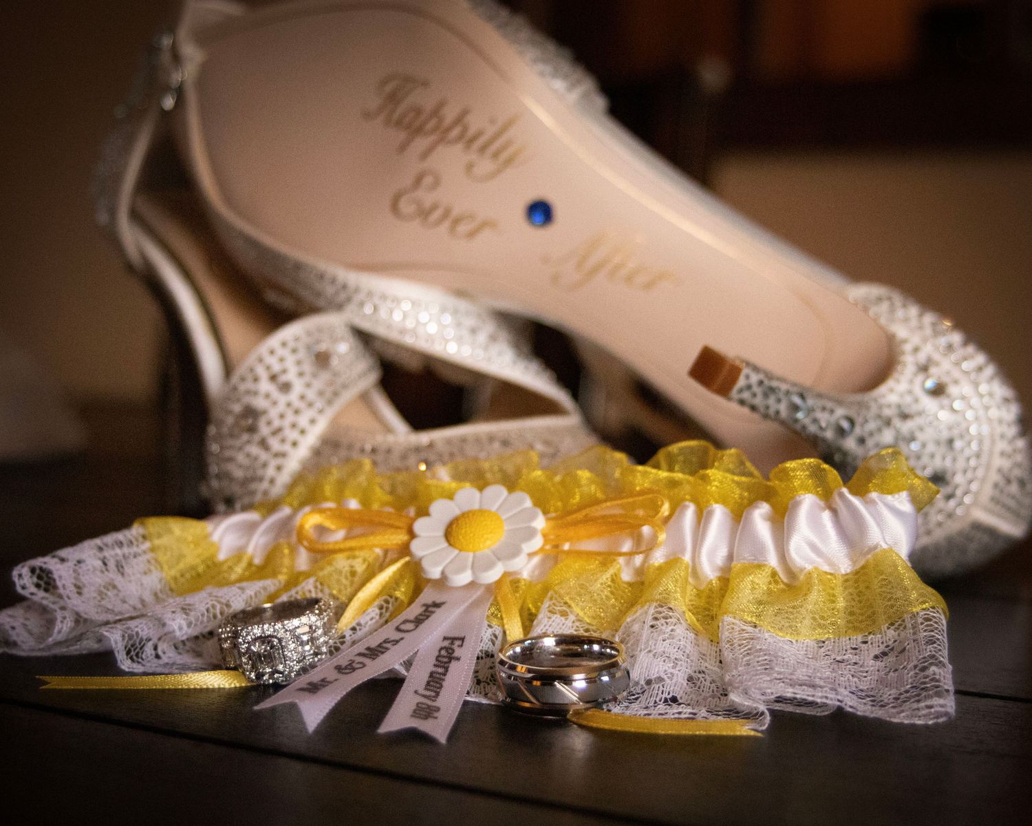 Wedding accessories featuring crystal-embellished bridal shoes and a yellow and white lace garter on dark surface.