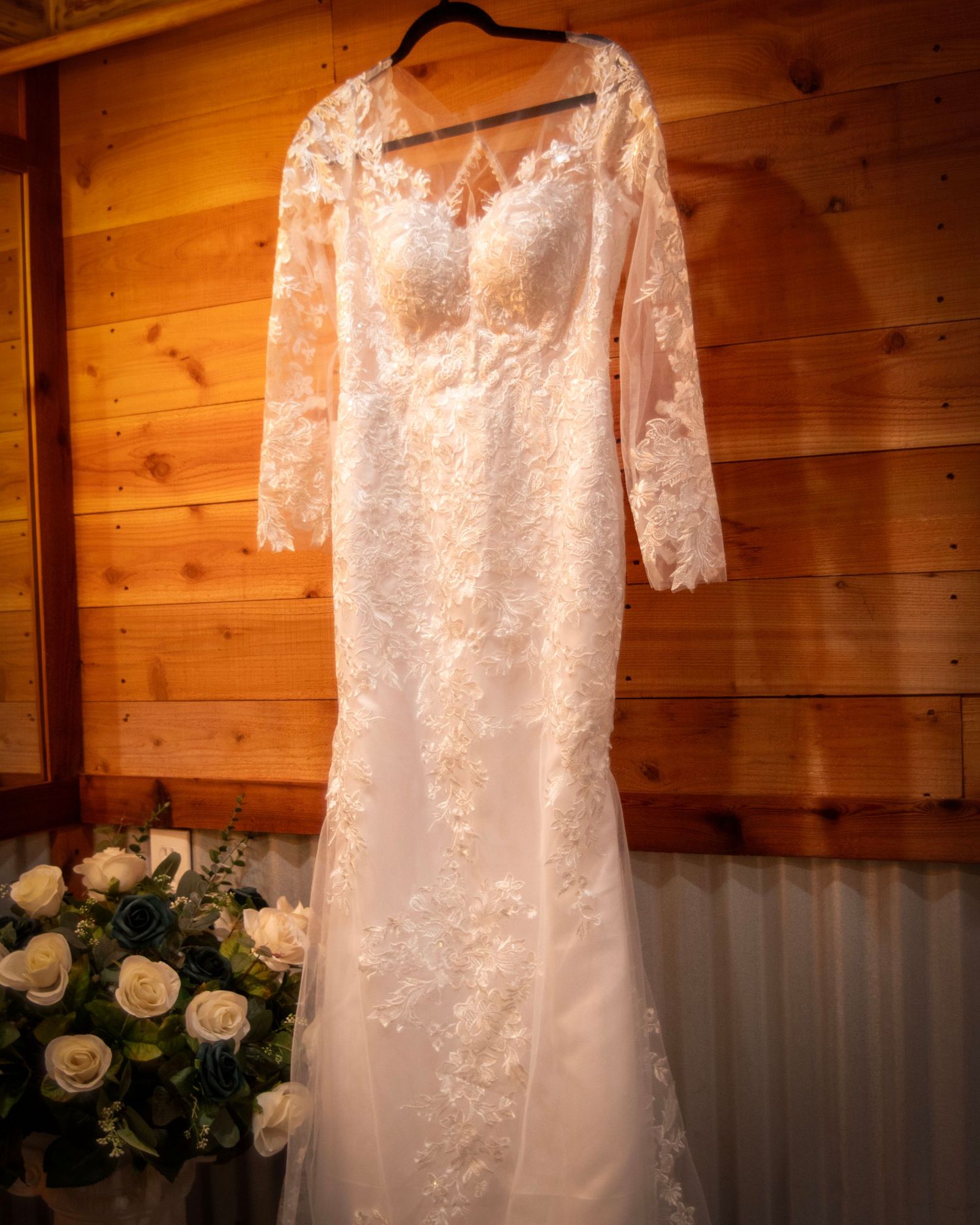 An elegant white lace wedding dress with long sleeves hangs against a wooden wall beside white rose bouquet decorations.