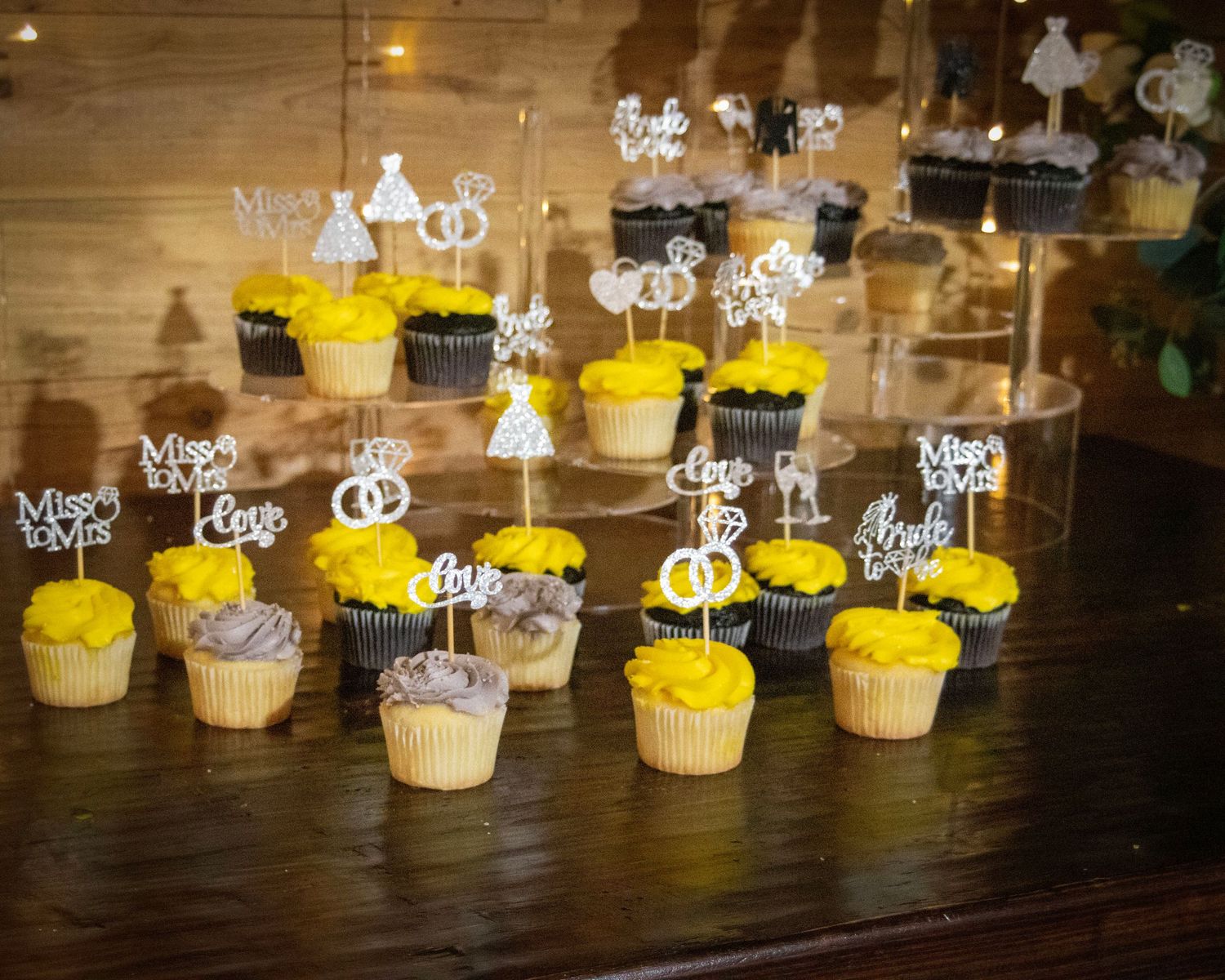Yellow and white frosted cupcakes displayed with decorative toppers against a rustic wooden background with string lights.