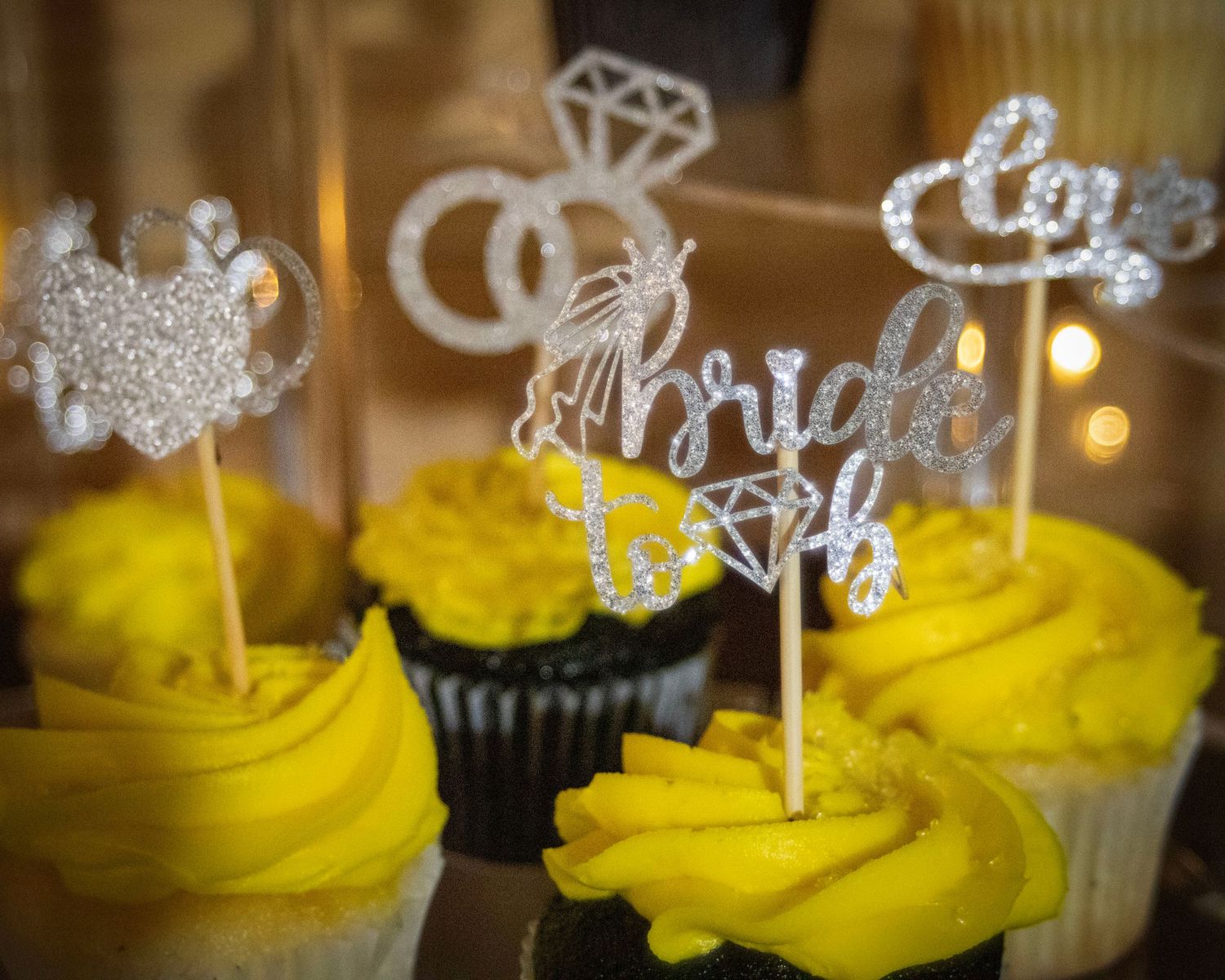 Chocolate cupcakes with bright yellow frosting and sparkling silver bridal-themed cake toppers.