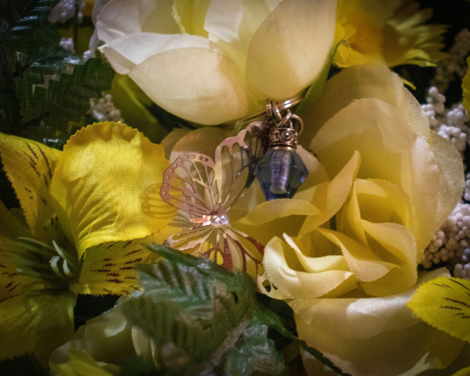 Close-up of a delicate butterfly pendant on a blue vial nestled among yellow and cream roses.