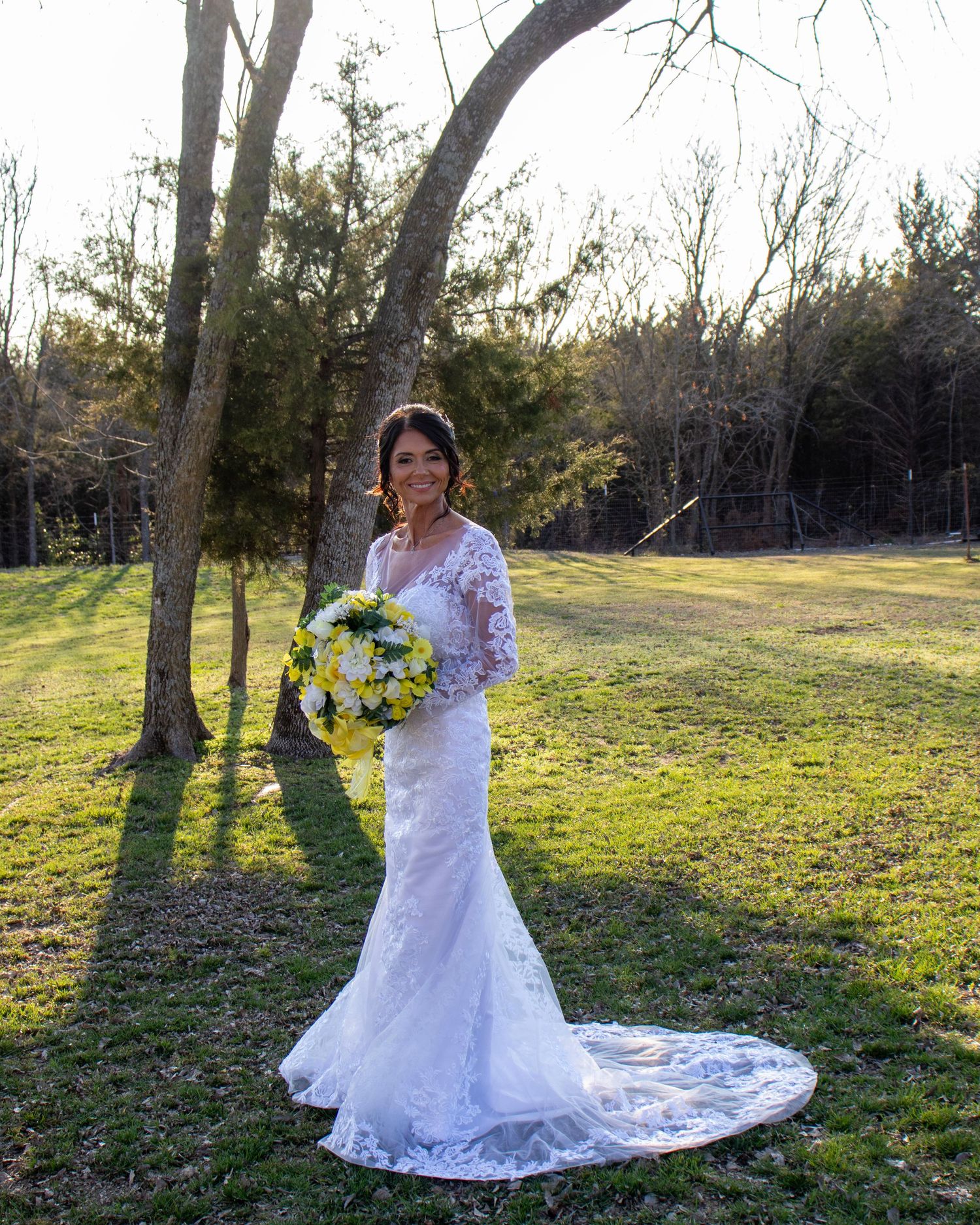 A bride in a white lace gown with long train holds a yellow and white bouquet while standing in a sunlit field at dusk.