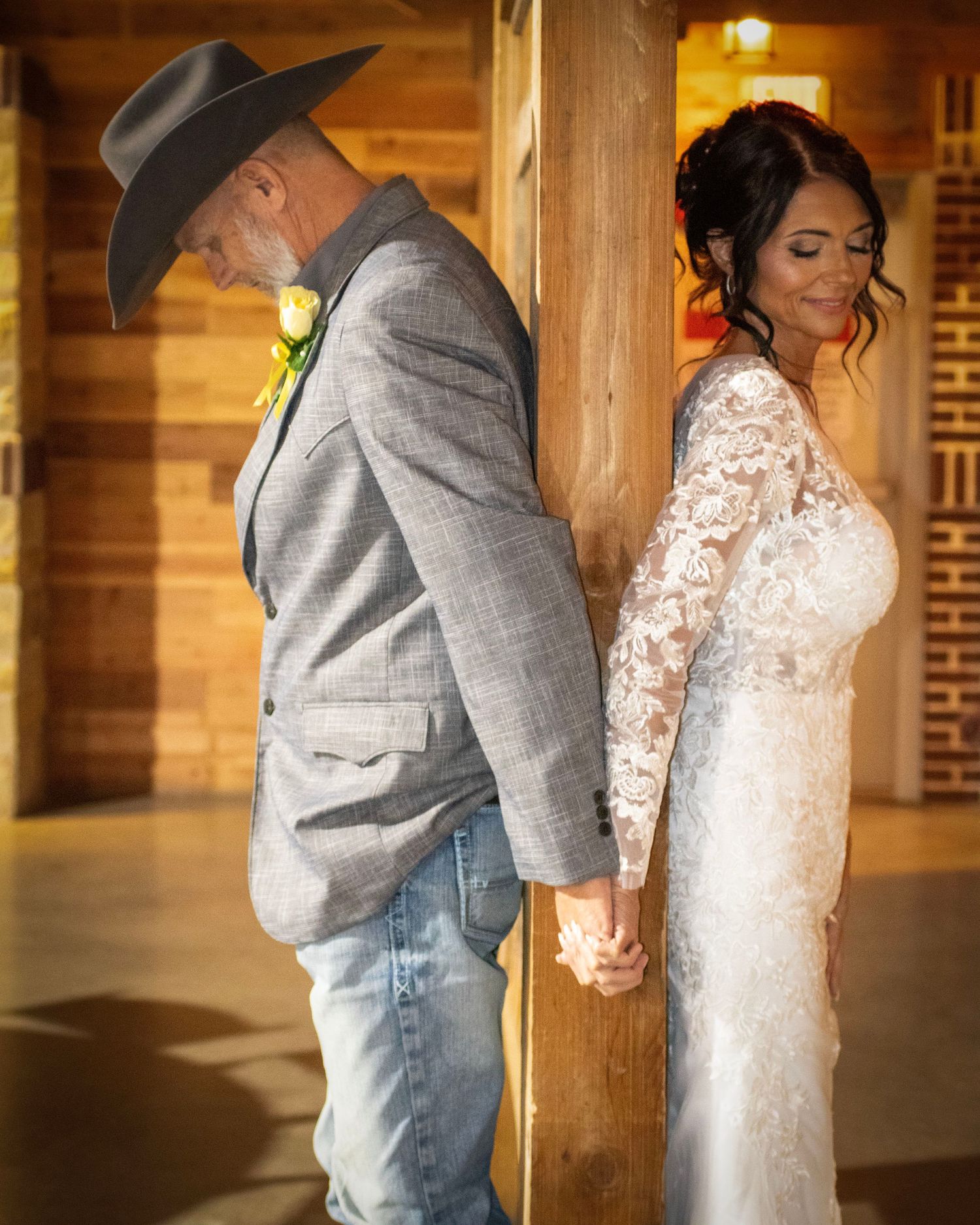 Bride in lace gown and groom in cowboy hat share a tender moment against wooden post at country wedding.