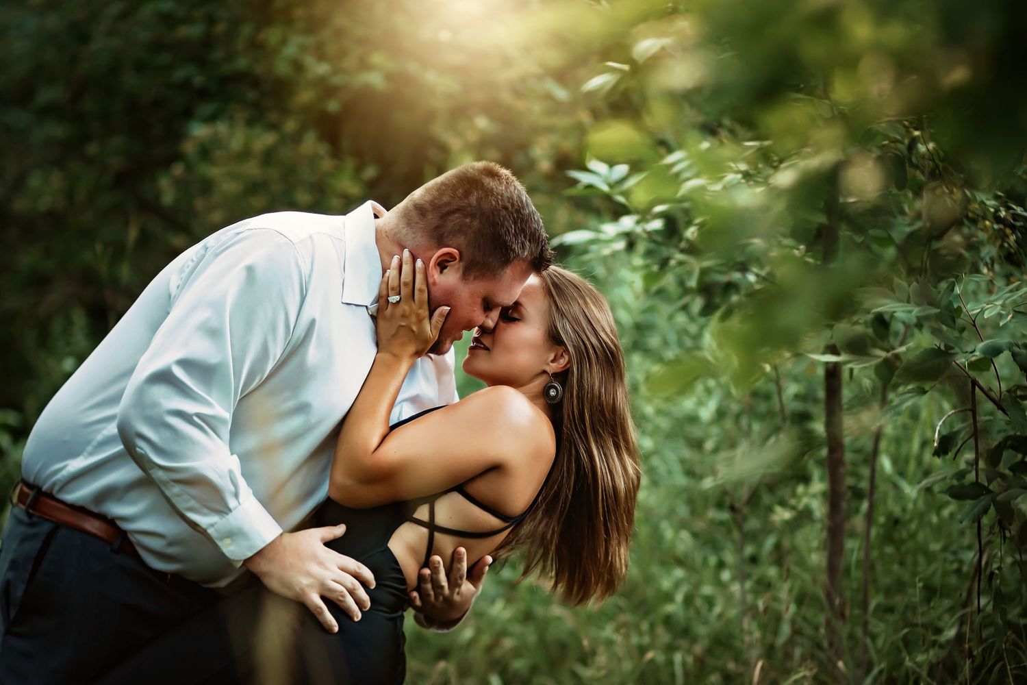 Couple shares a passionate kiss outdoors, surrounded by vibrant green foliage in a romantic natural setting.
