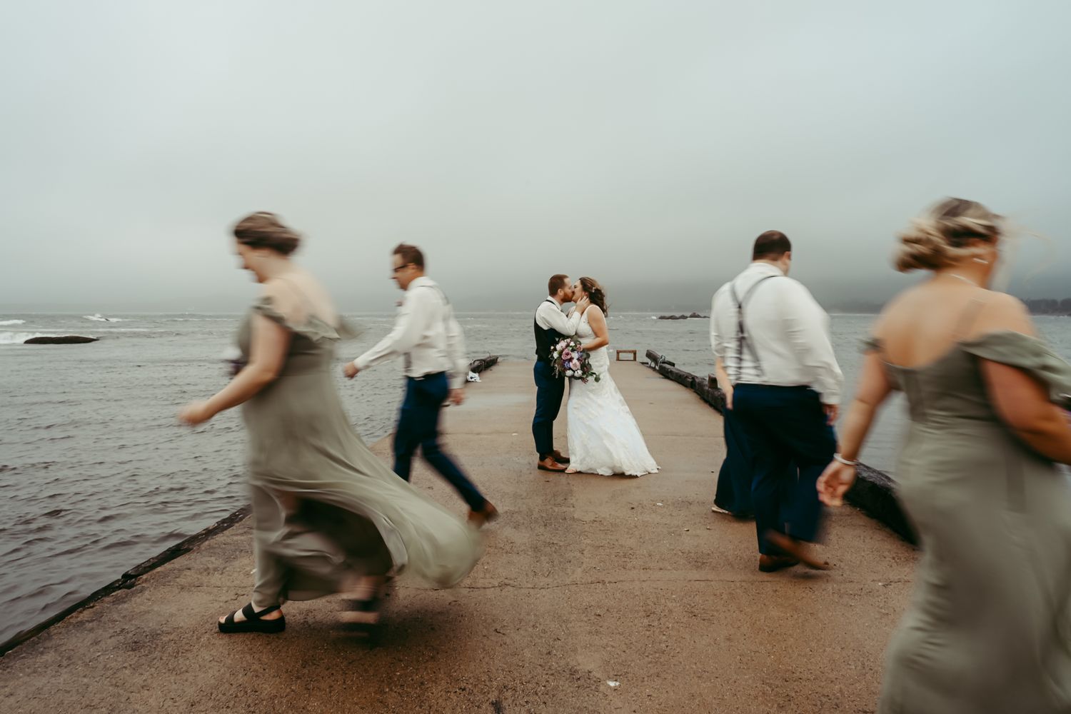 Wedding party runs and dances on a beach during overcast weather.