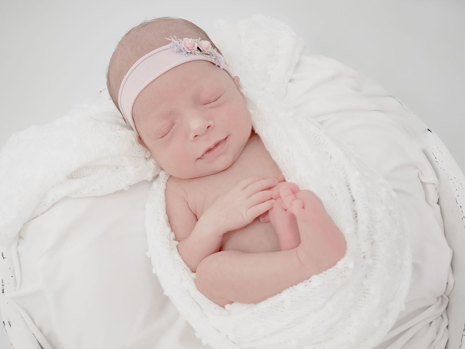 A peaceful sleeping newborn wrapped in soft white fabric wearing a delicate pink headband rests serenely.