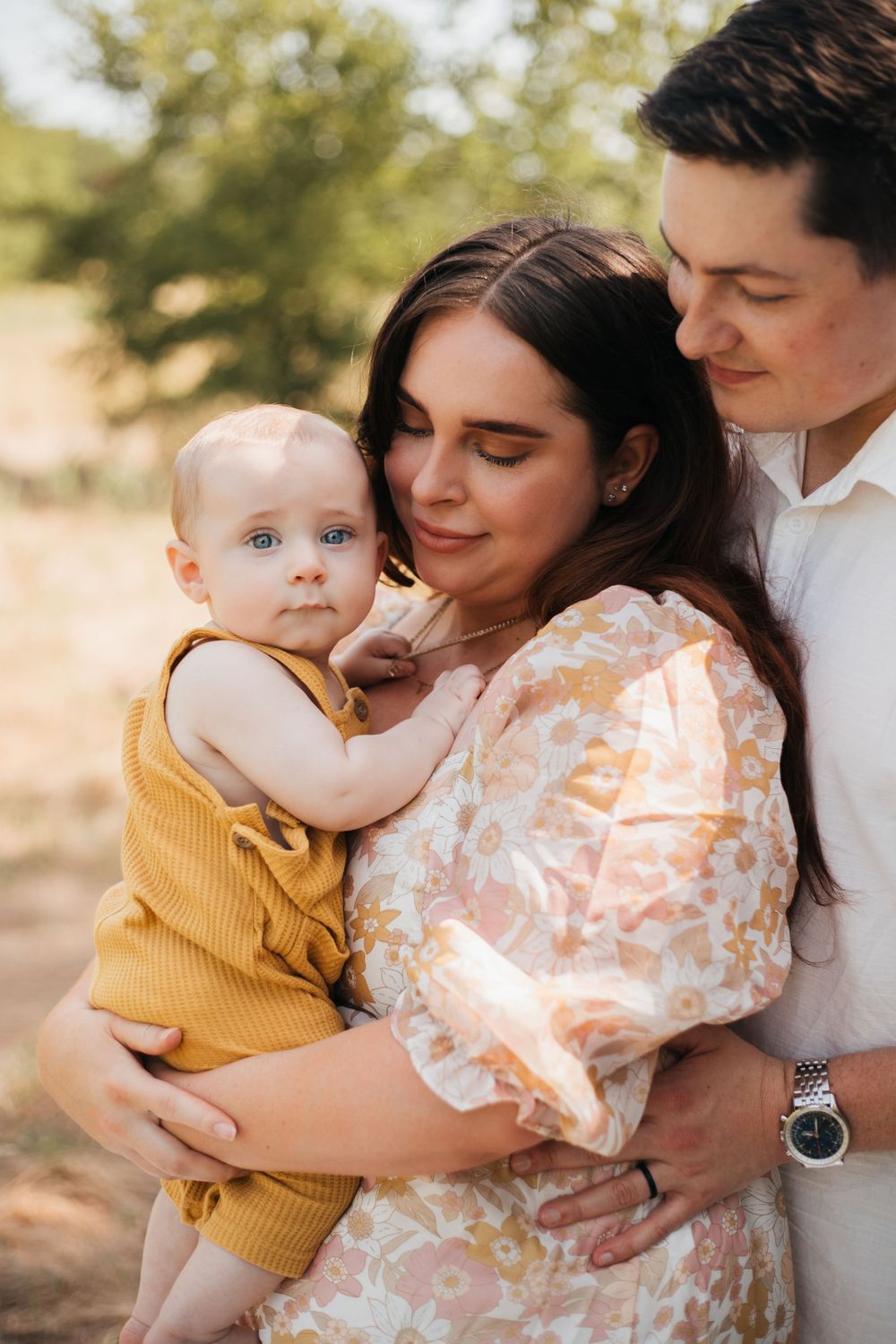 Intimate outdoor family portrait with baby in mustard overalls against natural background.