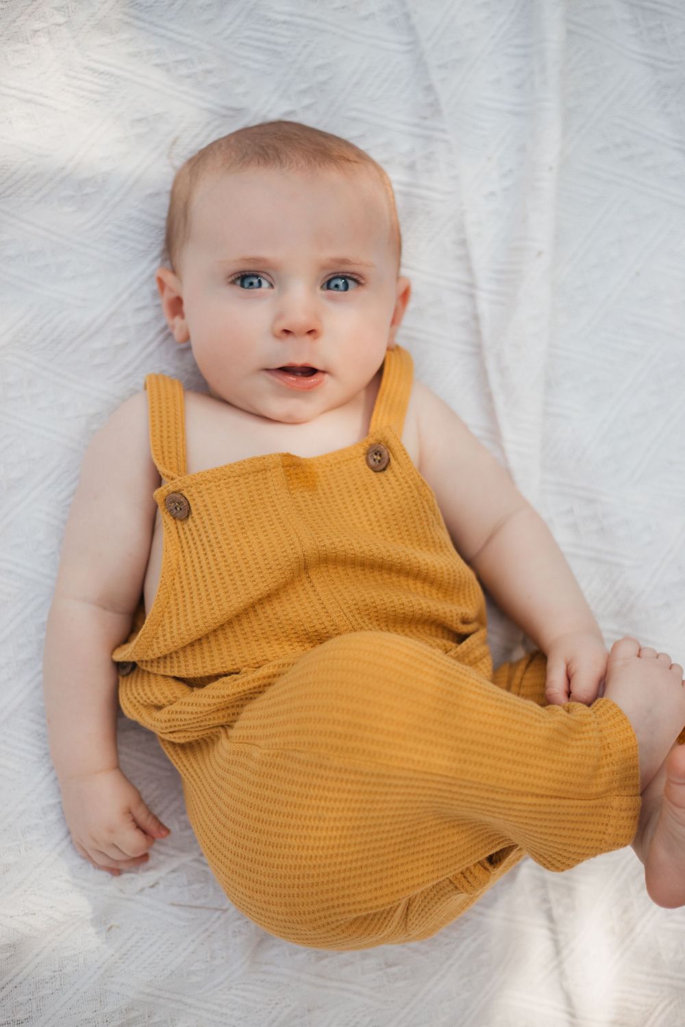 Baby wearing yellow ribbed overalls lying on white textured blanket.