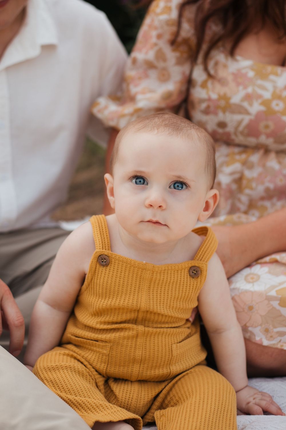 Baby in mustard yellow overalls with bright blue eyes sits on blanket outdoors.