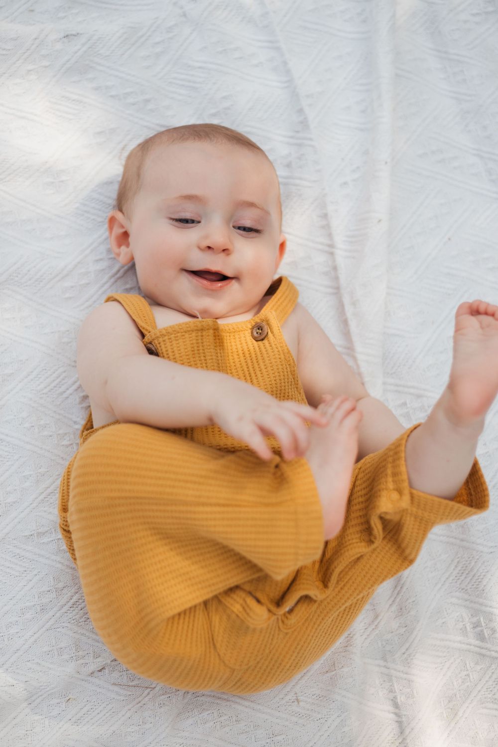 Happy baby in mustard romper playing on white blanket with natural lighting.