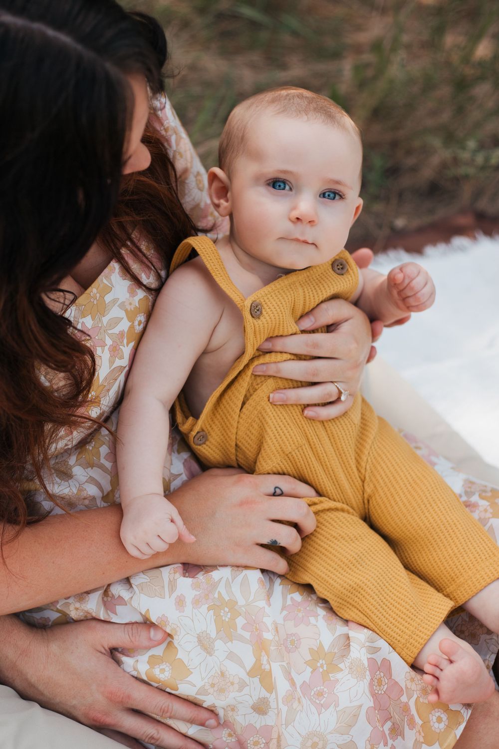 Baby wearing yellow knit overalls rests on floral dress during outdoor photoshoot.