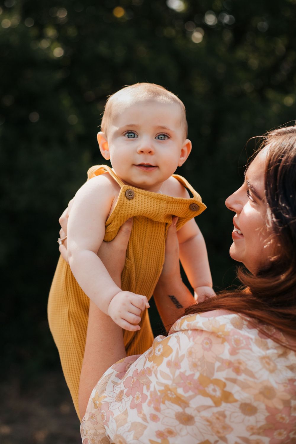 Baby in mustard yellow romper being held outdoors with soft natural lighting.
