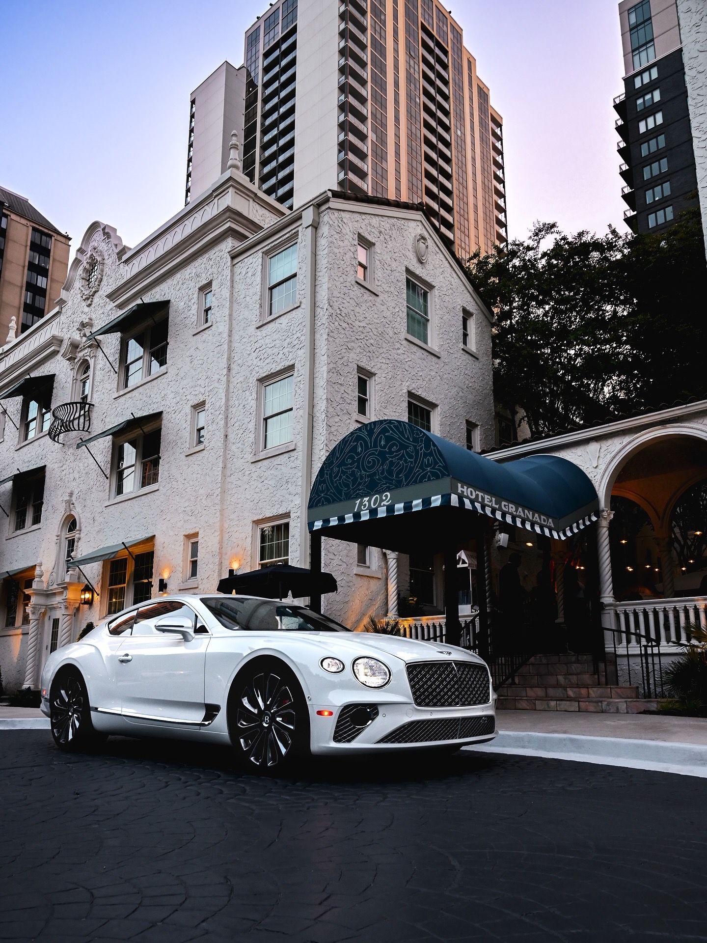A white Bentley Continental GT parked in front of a luxury apartment building with a classic architecture entrance canopy.