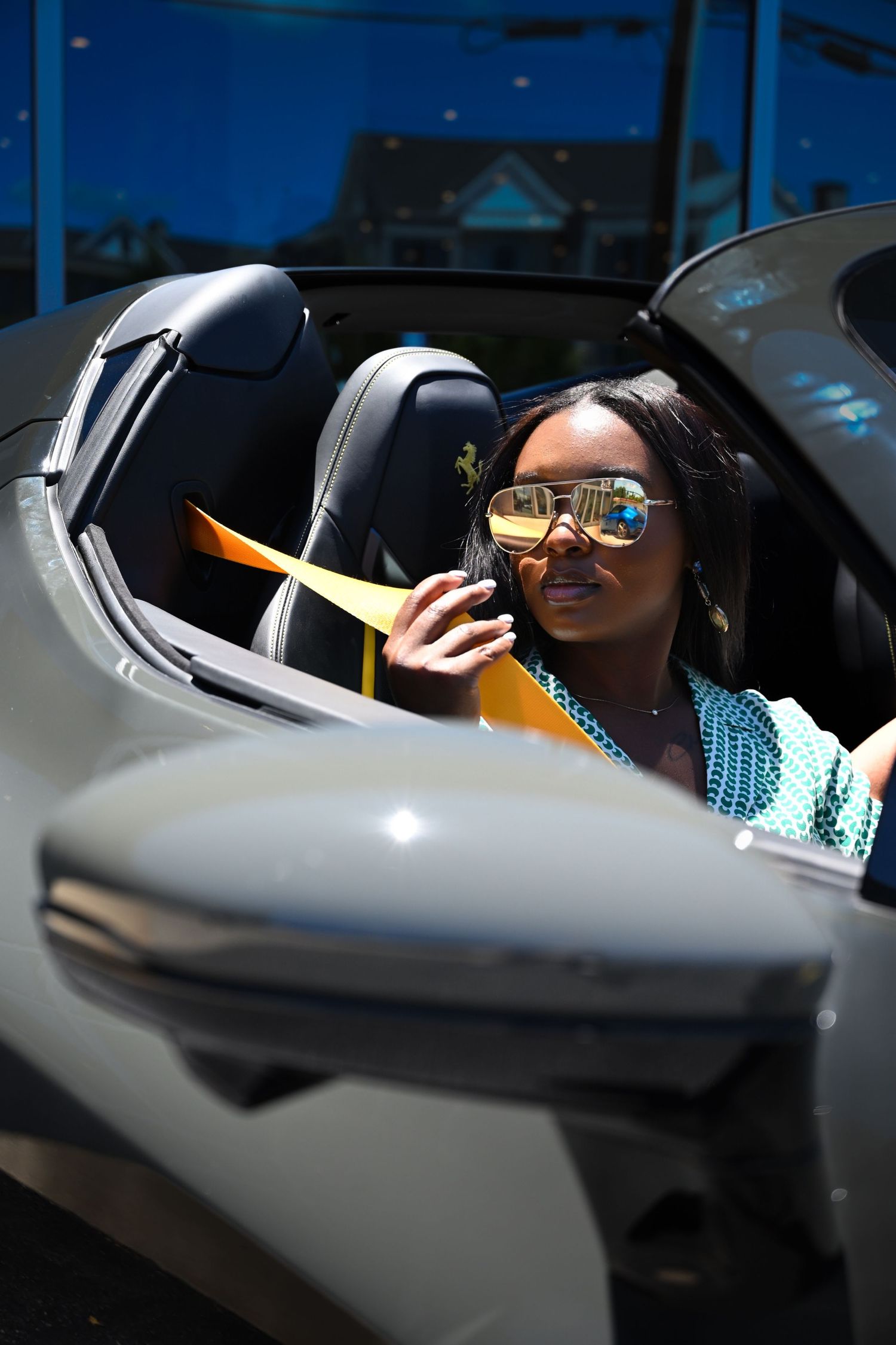 A person in a car wearing sunglasses and a blue top, viewed through the driver's side window with dramatic lighting.