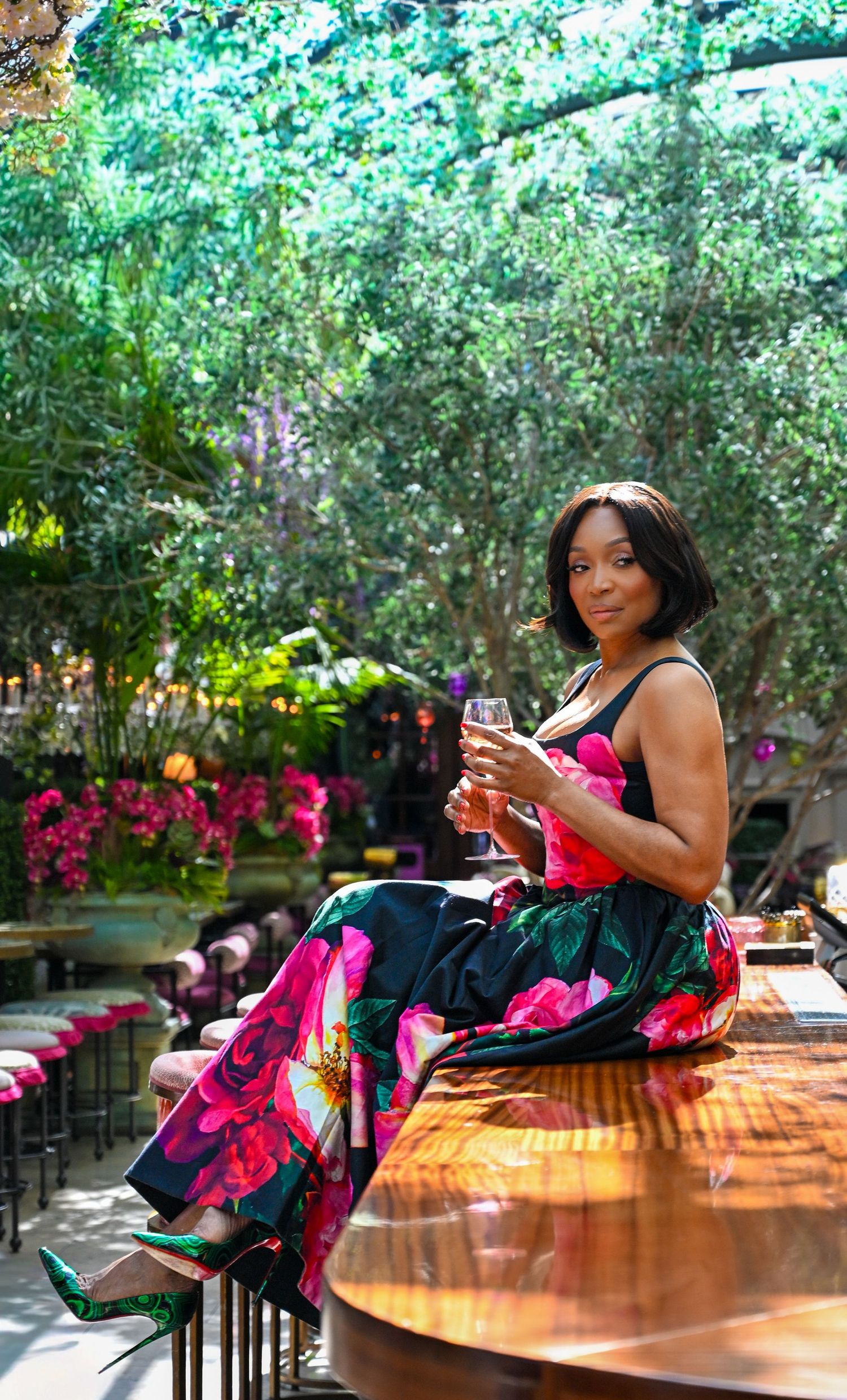 Woman in black dress with pink ruffles poses on wooden surface in garden setting with colorful flowers.