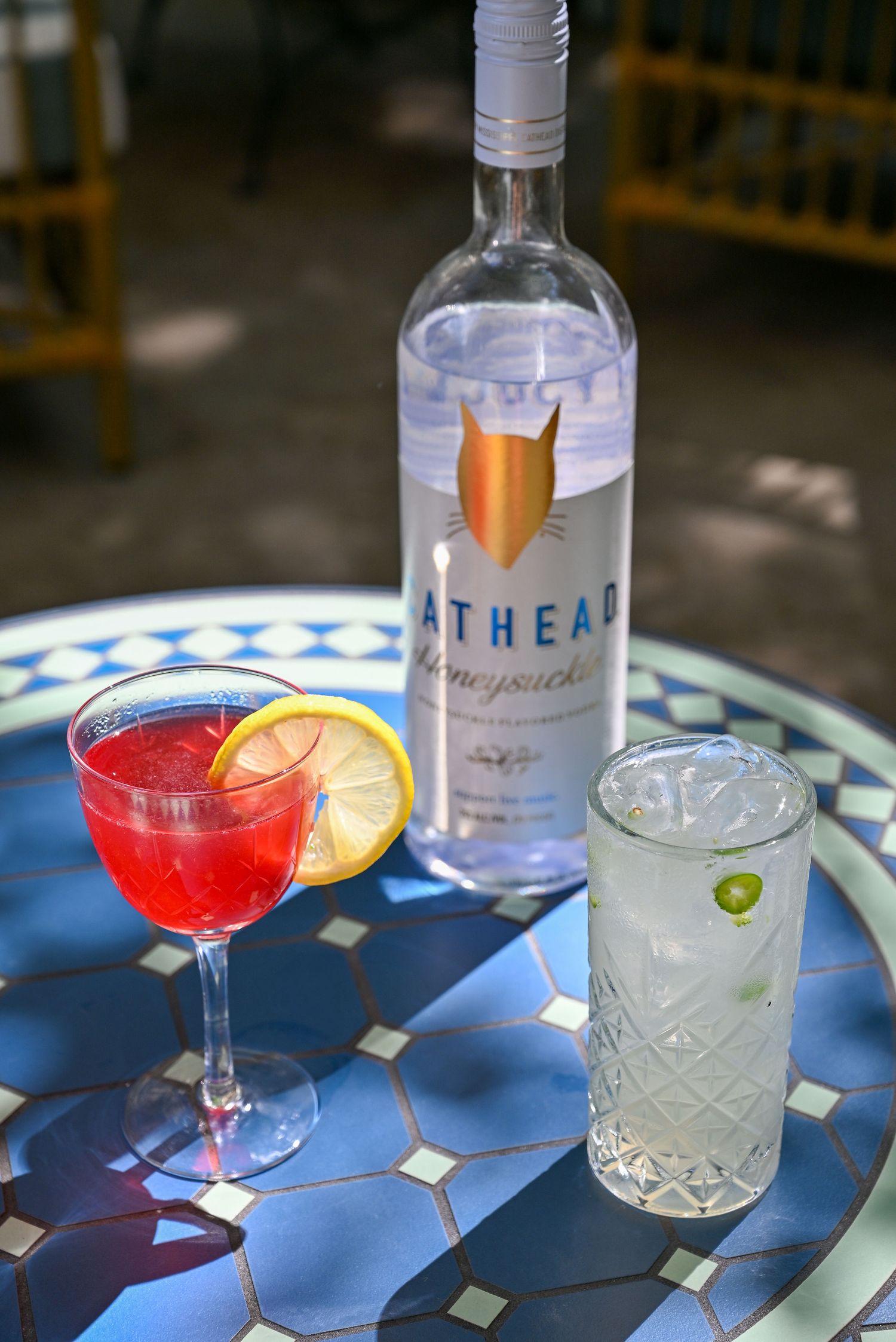 A bottle of Athean vodka with two cocktails on a blue mosaic table, one red with lemon and one clear in a crystal glass.