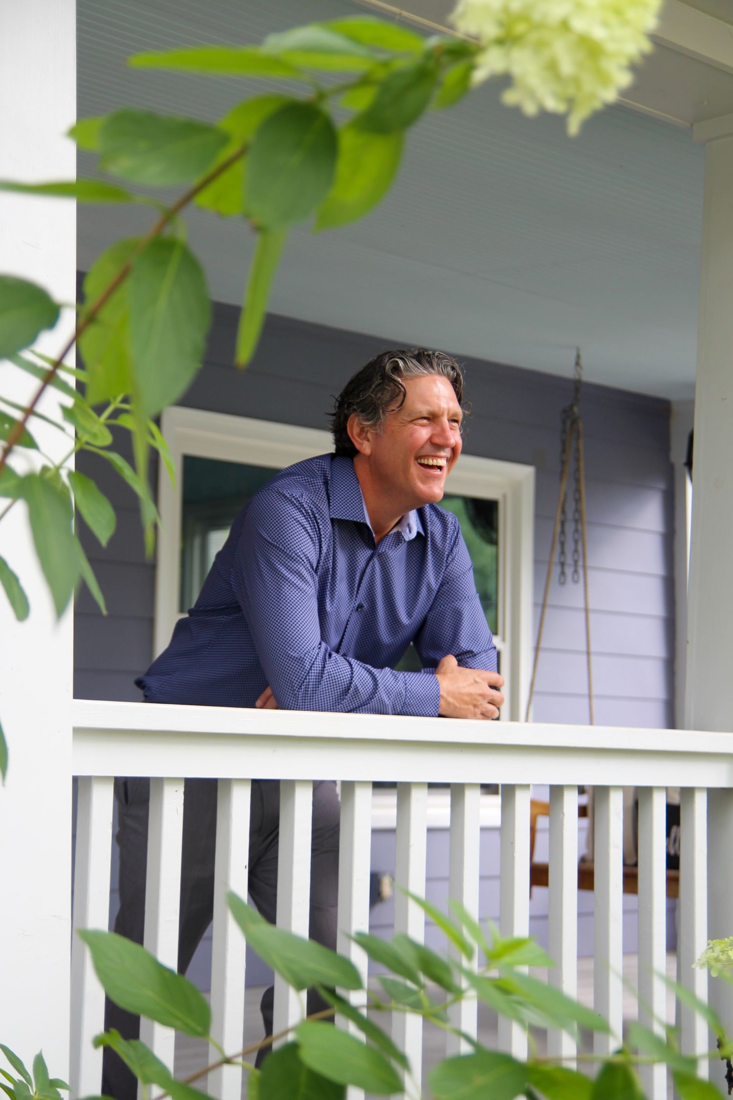 Person smiling while relaxing on a front porch with green foliage in the foreground.
