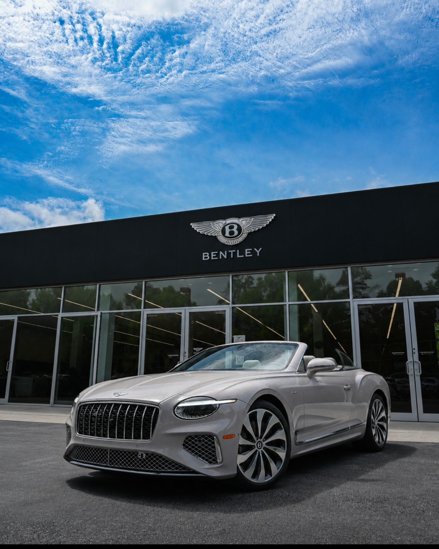 Silver Bentley convertible parked in front of a Bentley dealership under a blue sky with wispy clouds.