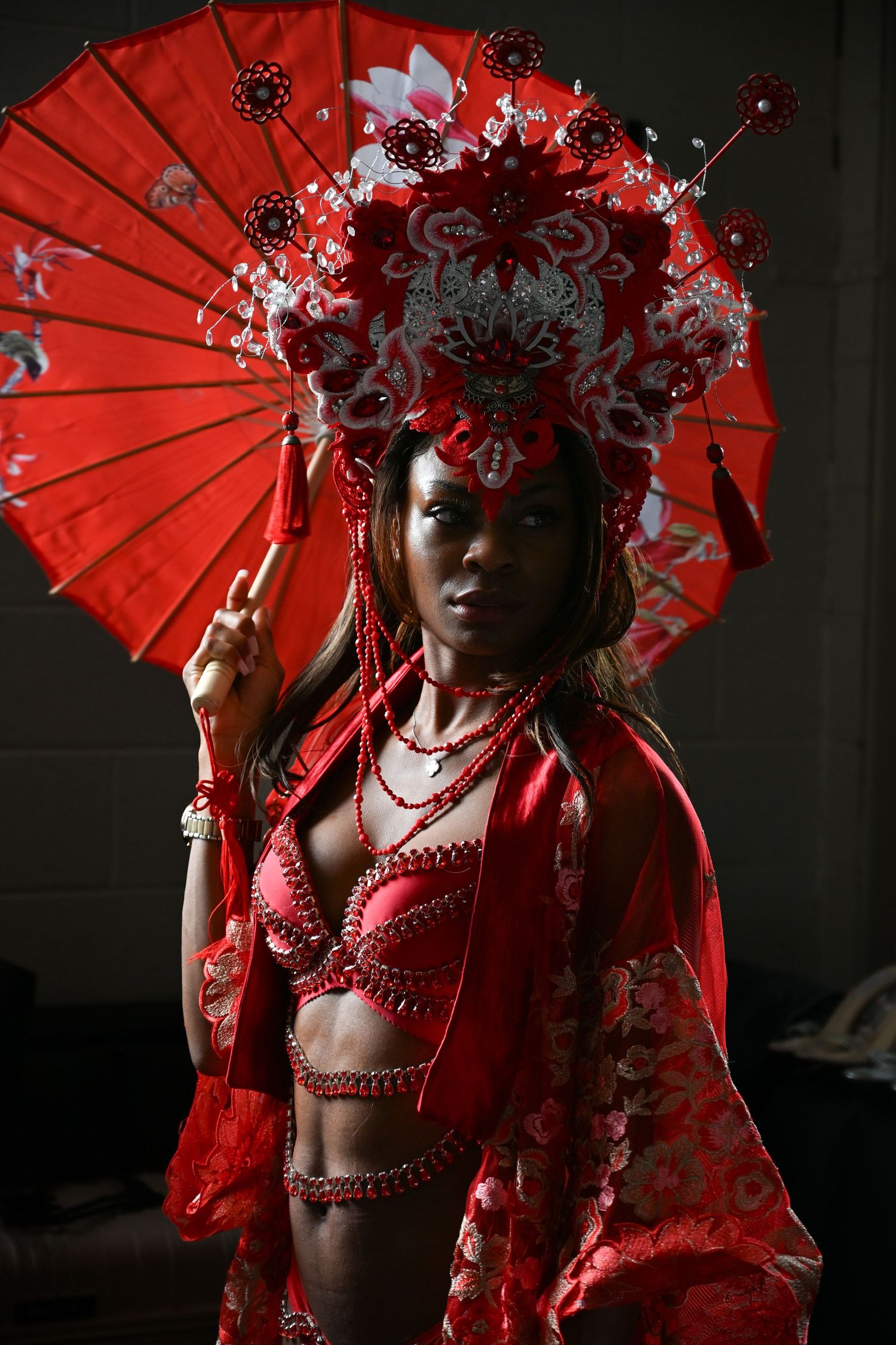 A figure in an ornate red traditional Asian-inspired costume with headdress and parasol poses dramatically against dark background.