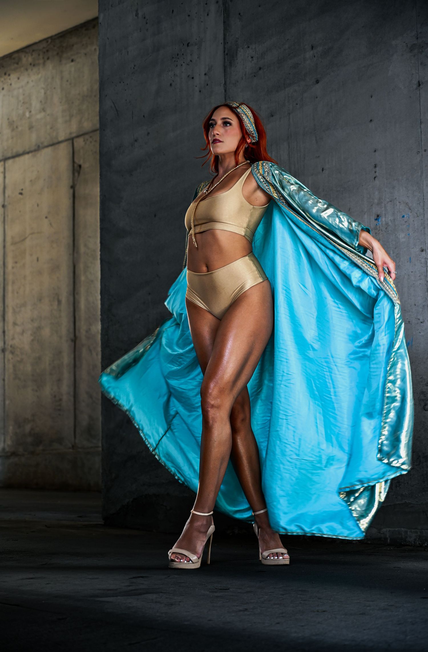 Fashion model poses dramatically in gold bikini and flowing turquoise silk robe against concrete wall backdrop.