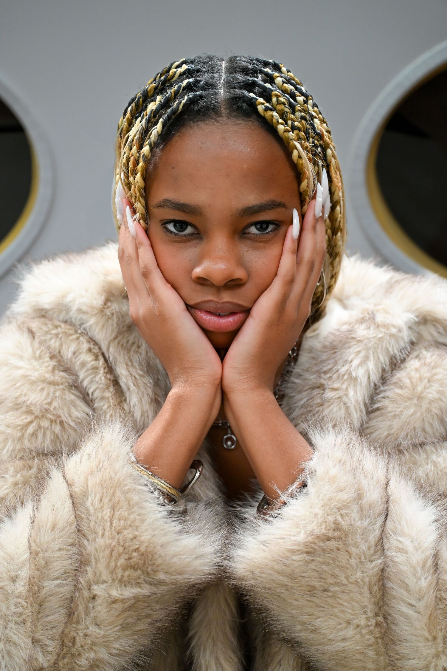 Fashion portrait of a model in a beige fur coat with blonde braids posing with hands on face against gray background.