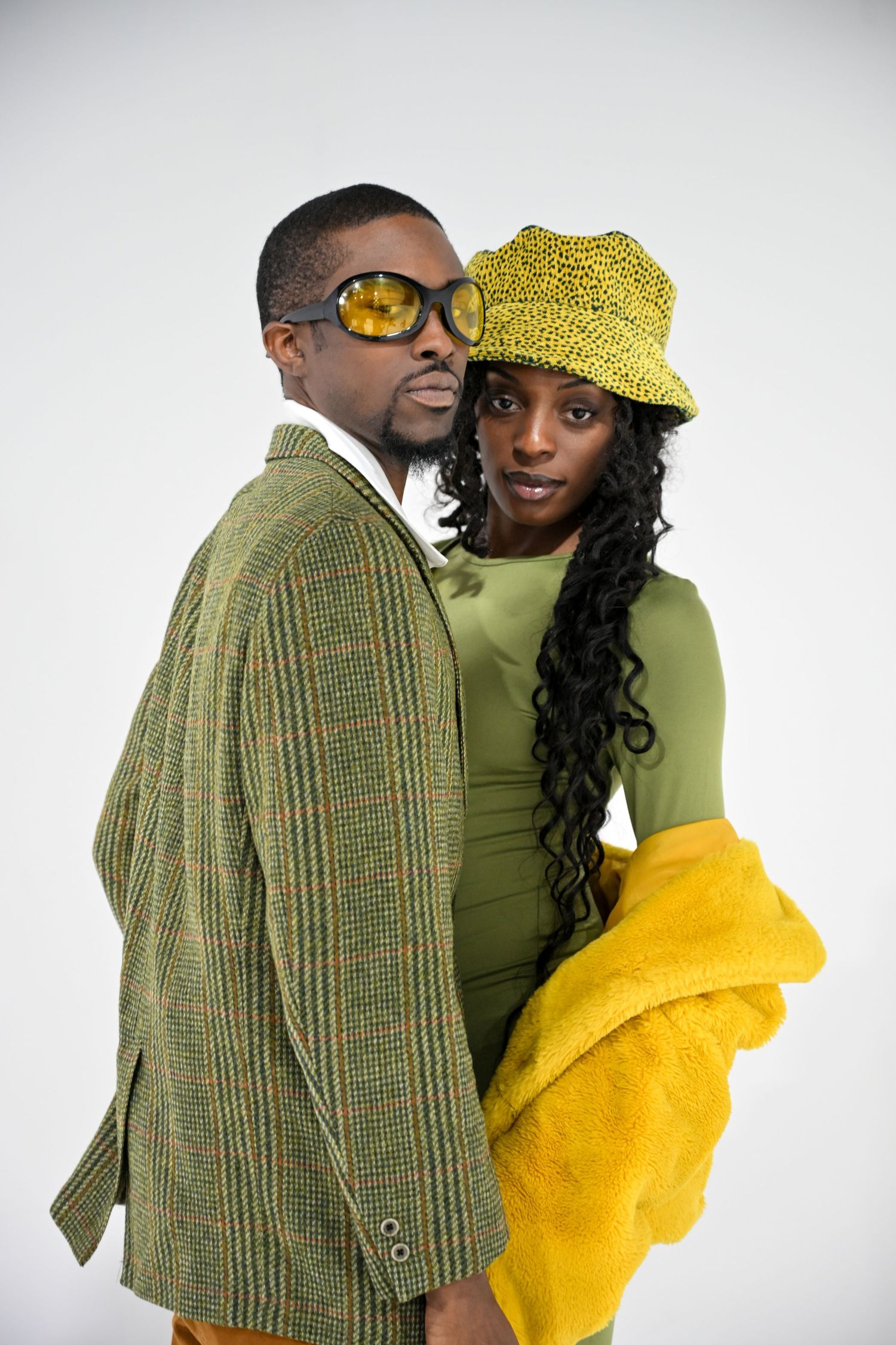 A couple embraces while wearing contrasting outfits of green tweed jacket, lime turtleneck, and yellow accessories against white.