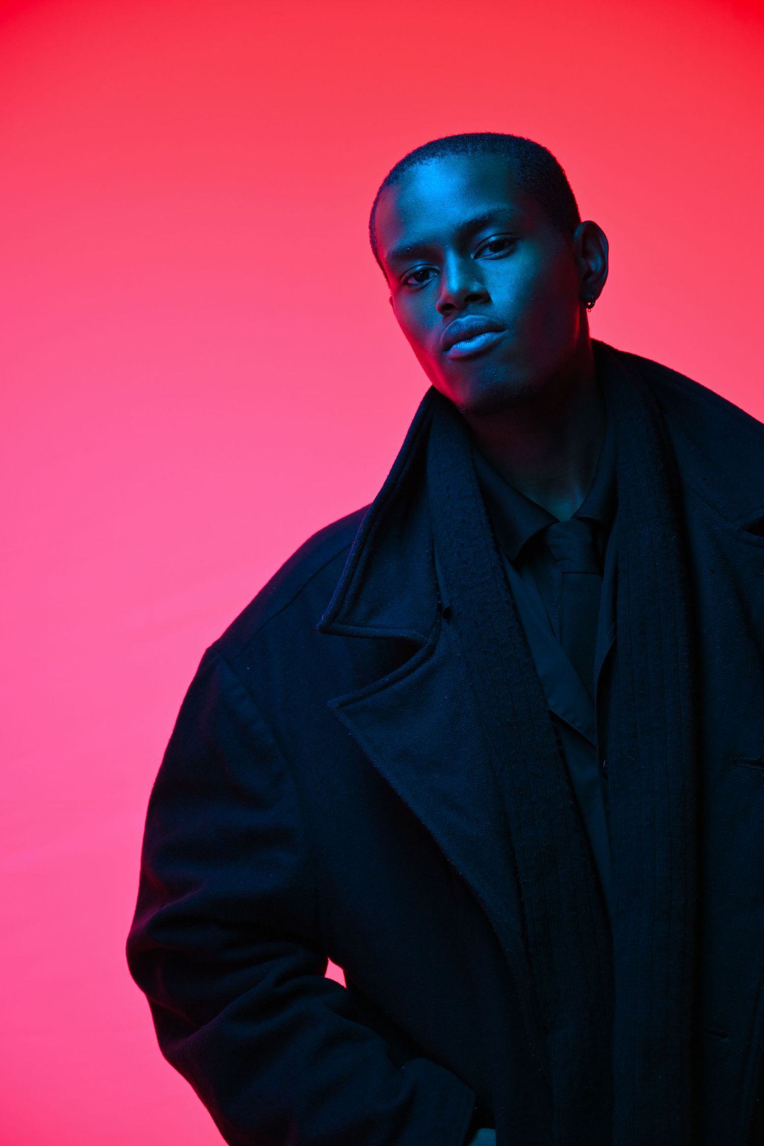 Artistic portrait in neon lighting showing a person in dark coat against vibrant pink background.