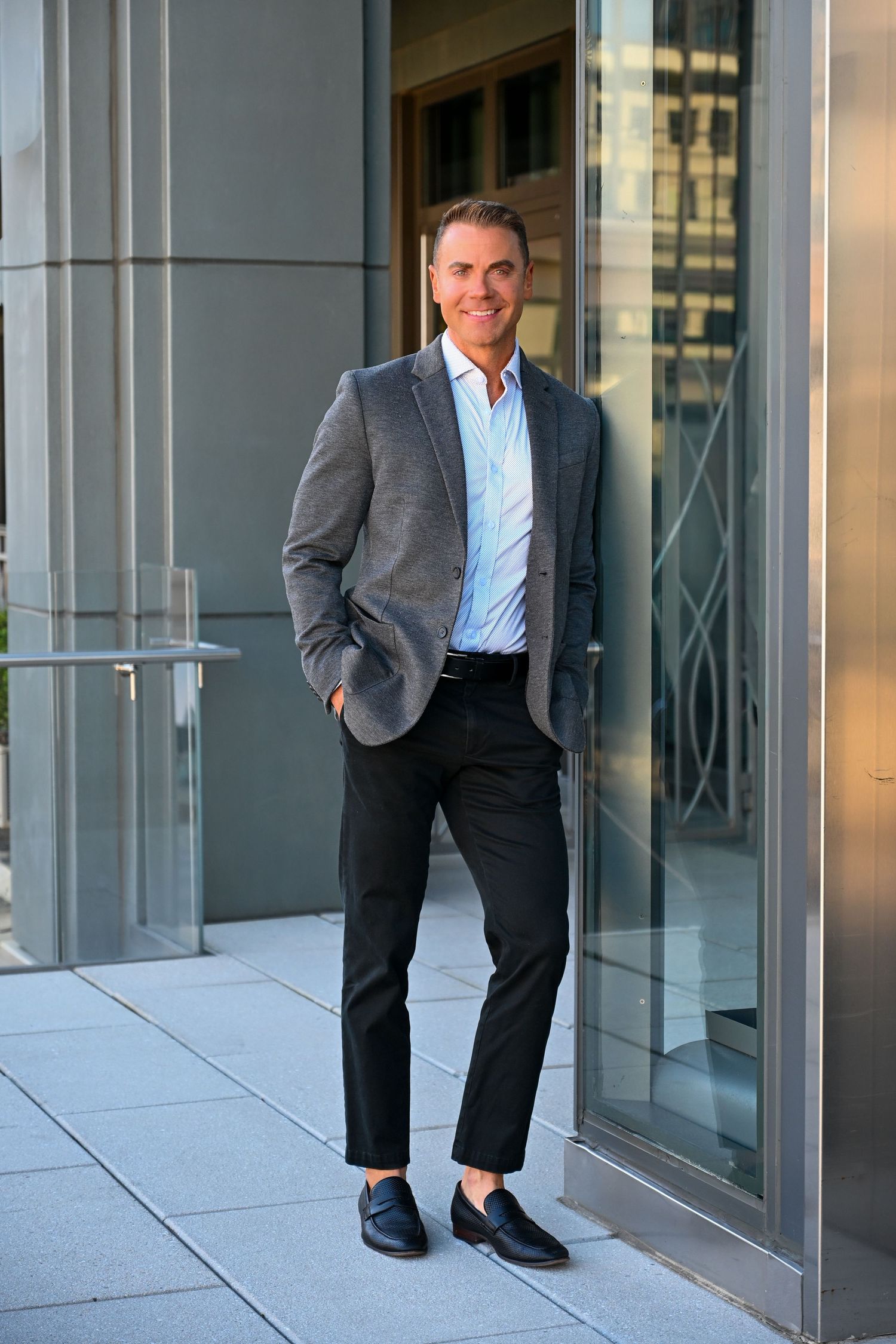 Professional in a dark suit and light blue shirt poses confidently outside a modern office building entrance.
